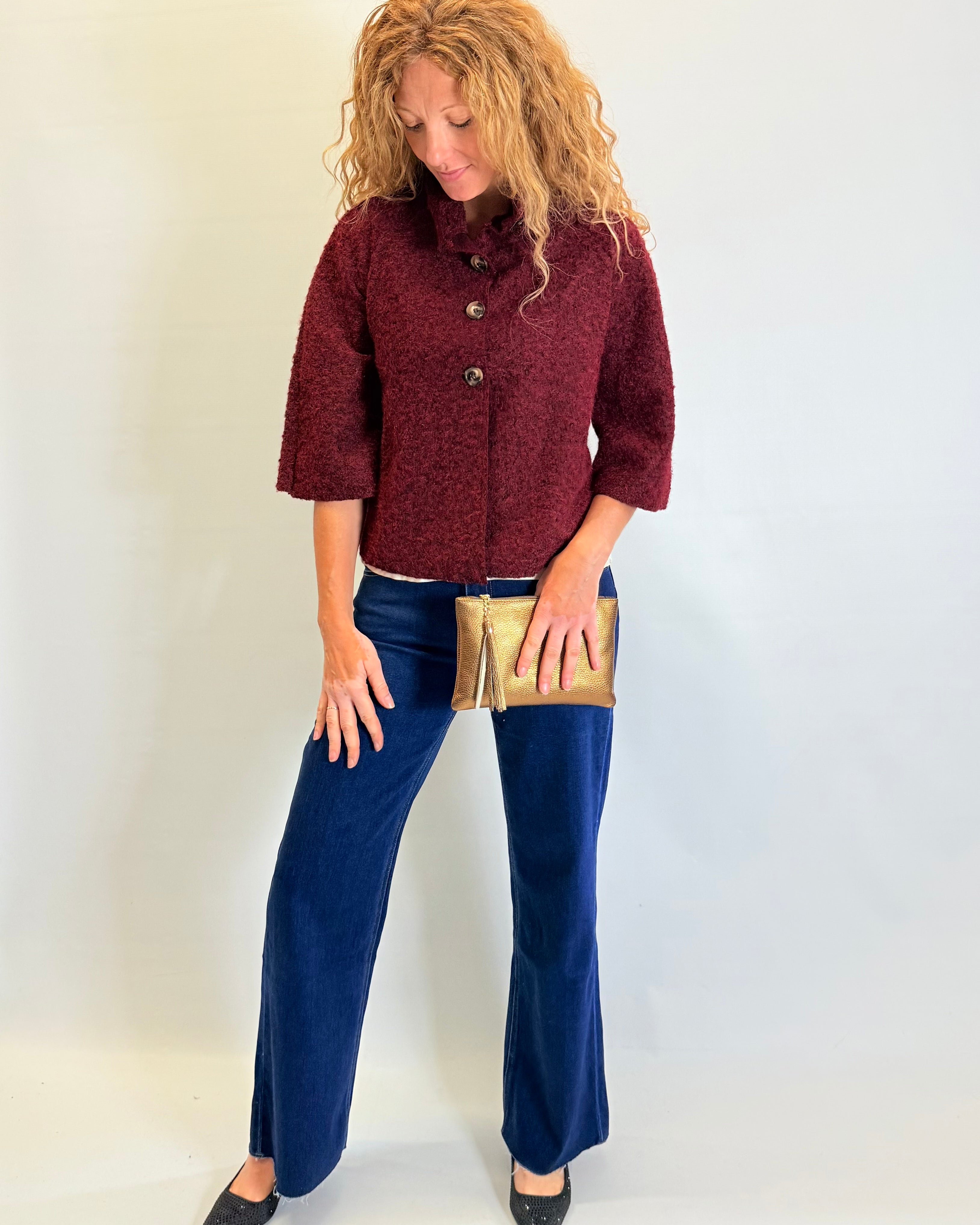 Woman wearing a burgundy jacket and blue pants holding a gold clutch against a white background