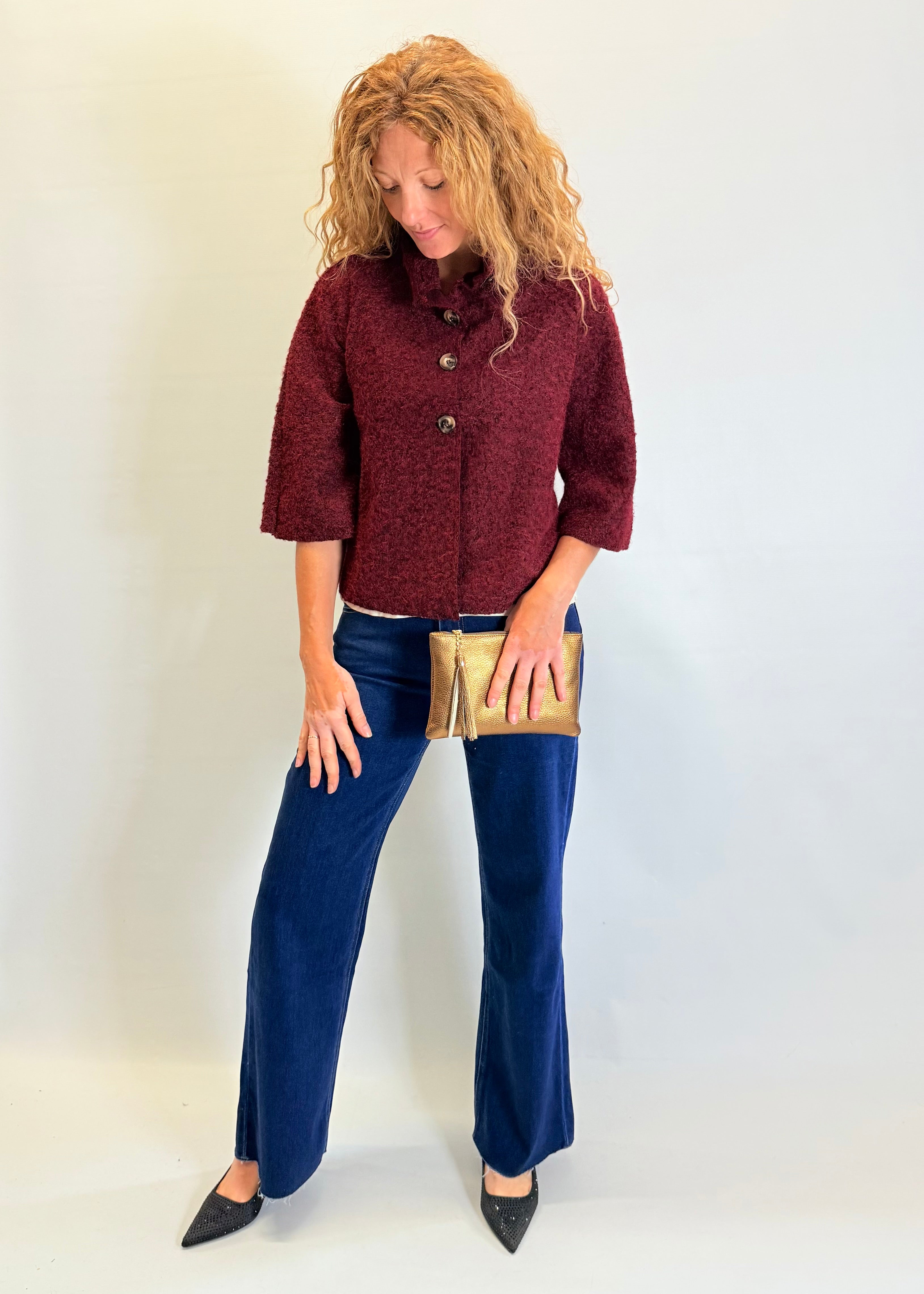 Woman wearing a burgundy jacket and blue pants holding a gold clutch against a white background