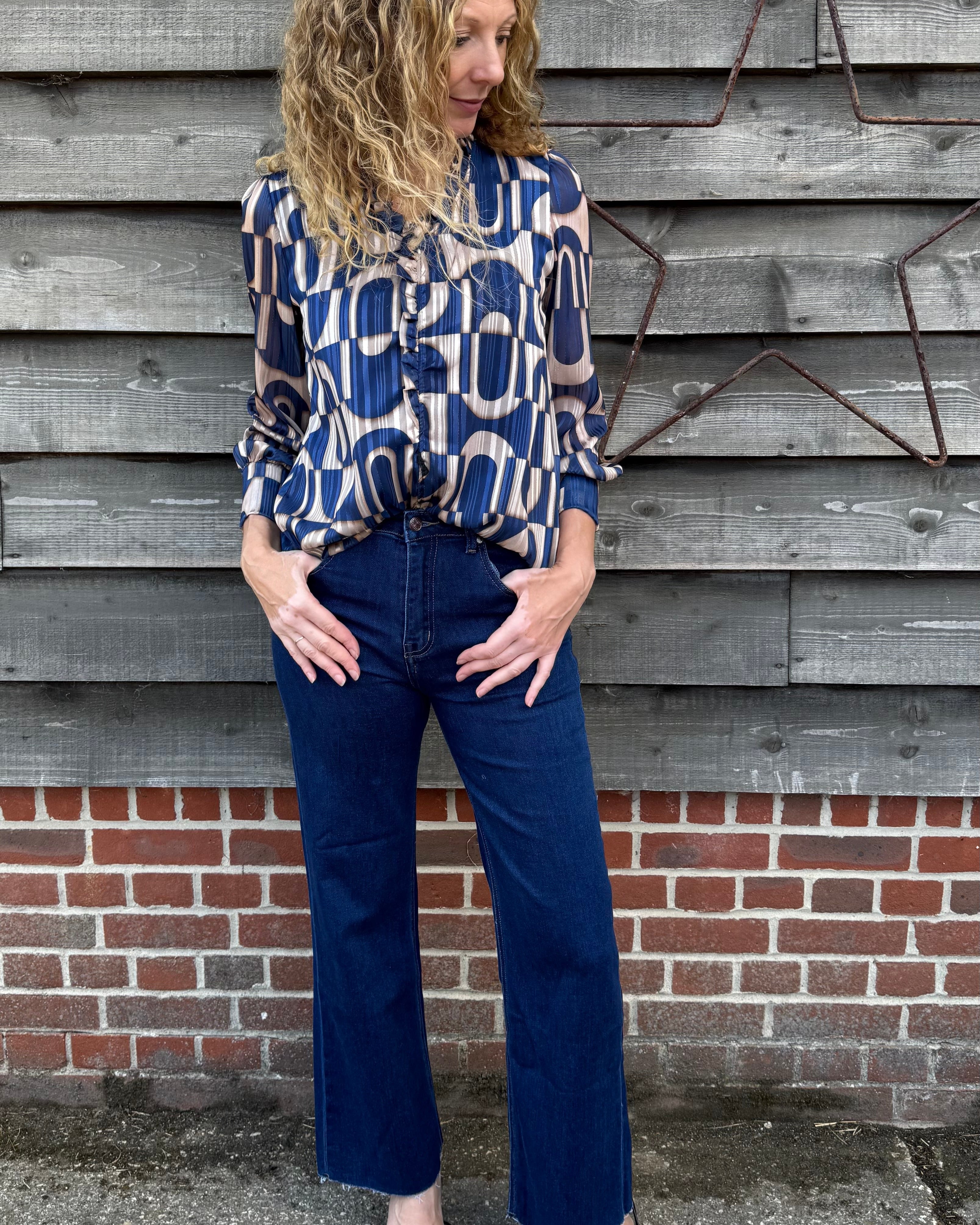 Woman wearing a patterned blouse and blue jeans standing against a wooden wall.