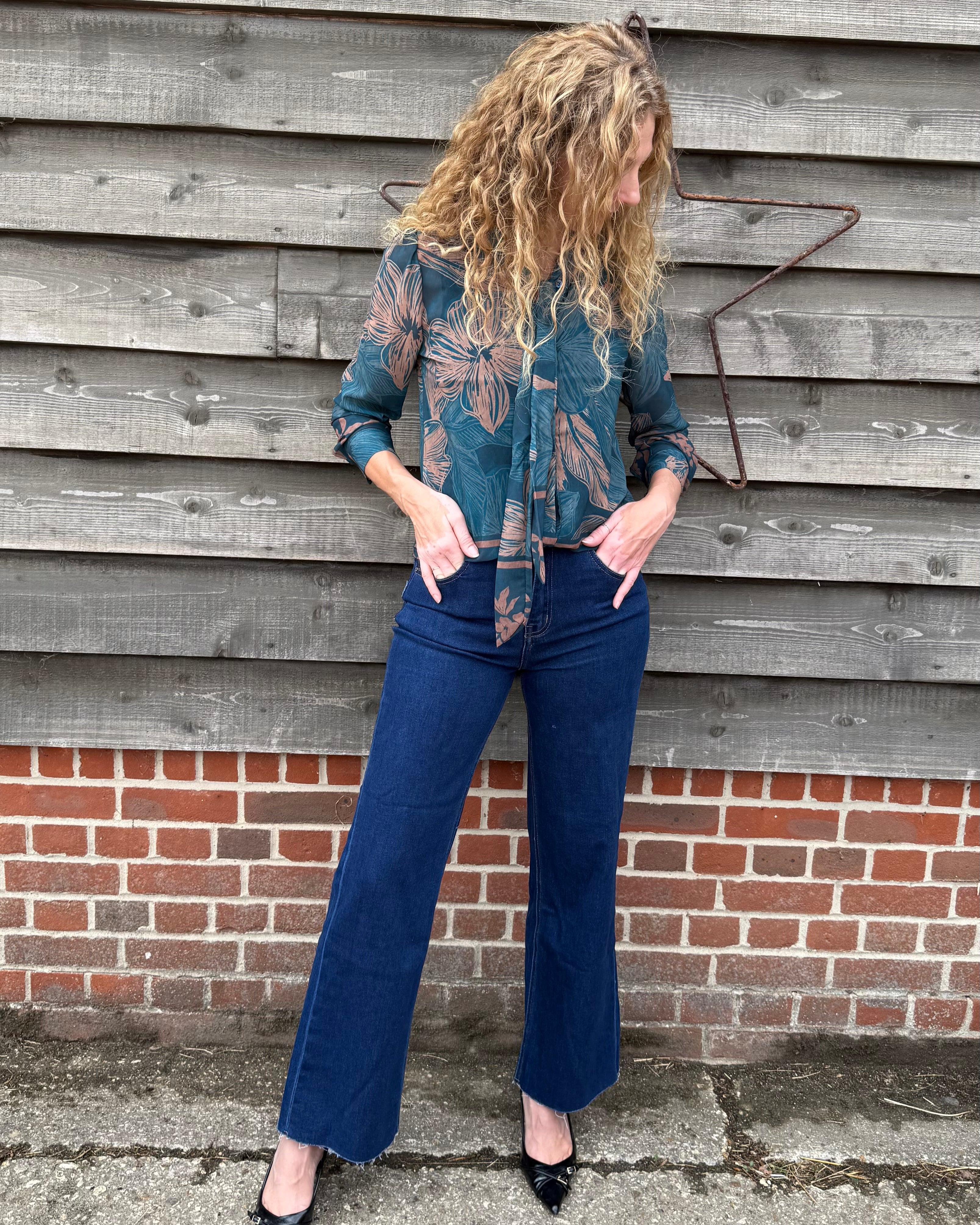 Person wearing a patterned shirt and blue jeans standing against a wooden wall.