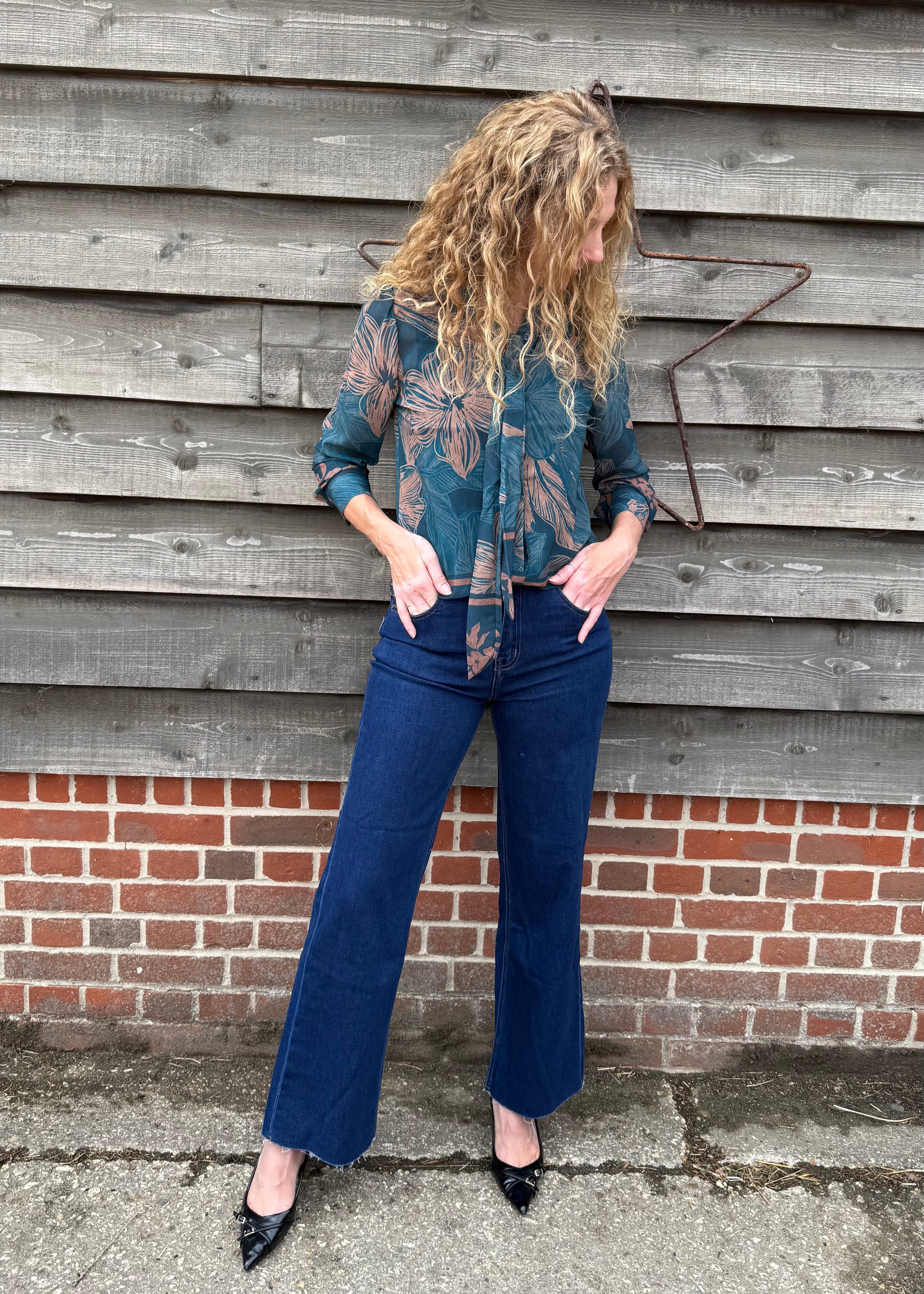 Person wearing a patterned shirt and blue jeans standing against a wooden wall.