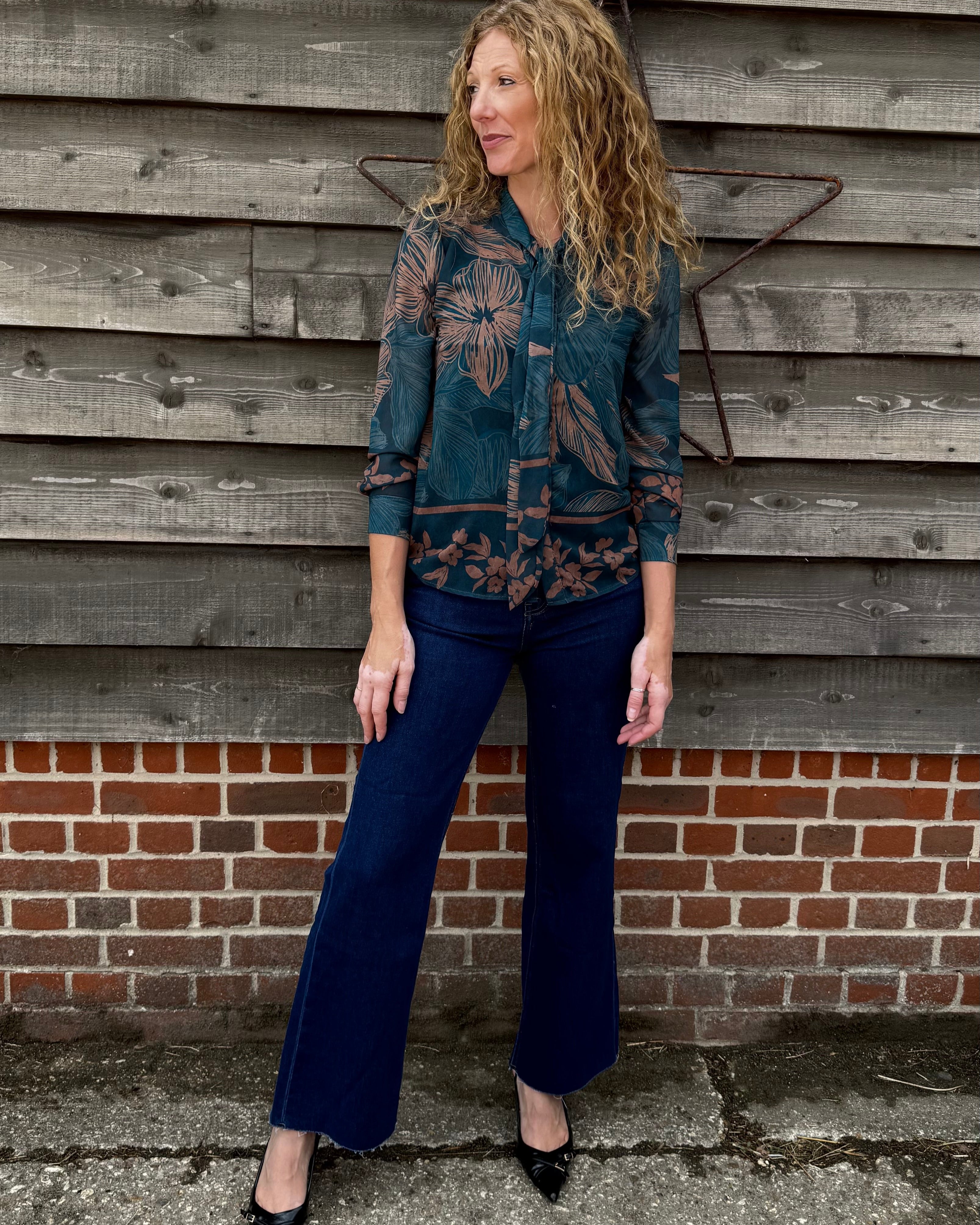Woman in a patterned shirt and blue jeans standing against a wooden wall.