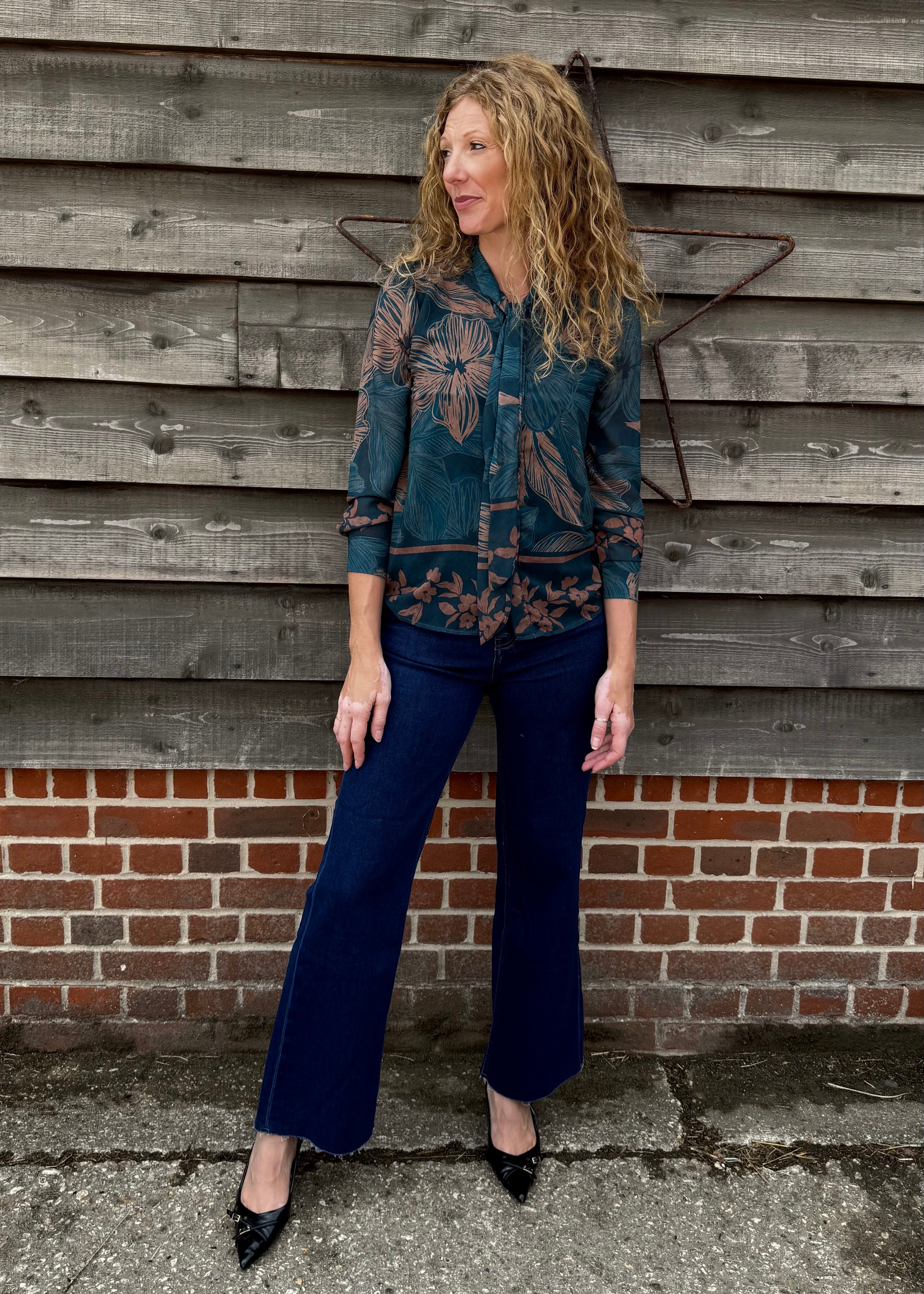 Woman in a patterned shirt and blue jeans standing against a wooden wall.