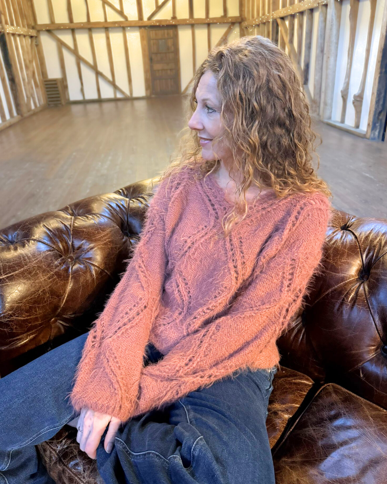 Woman wearing a pink sweater sitting on a brown leather couch in a rustic room.