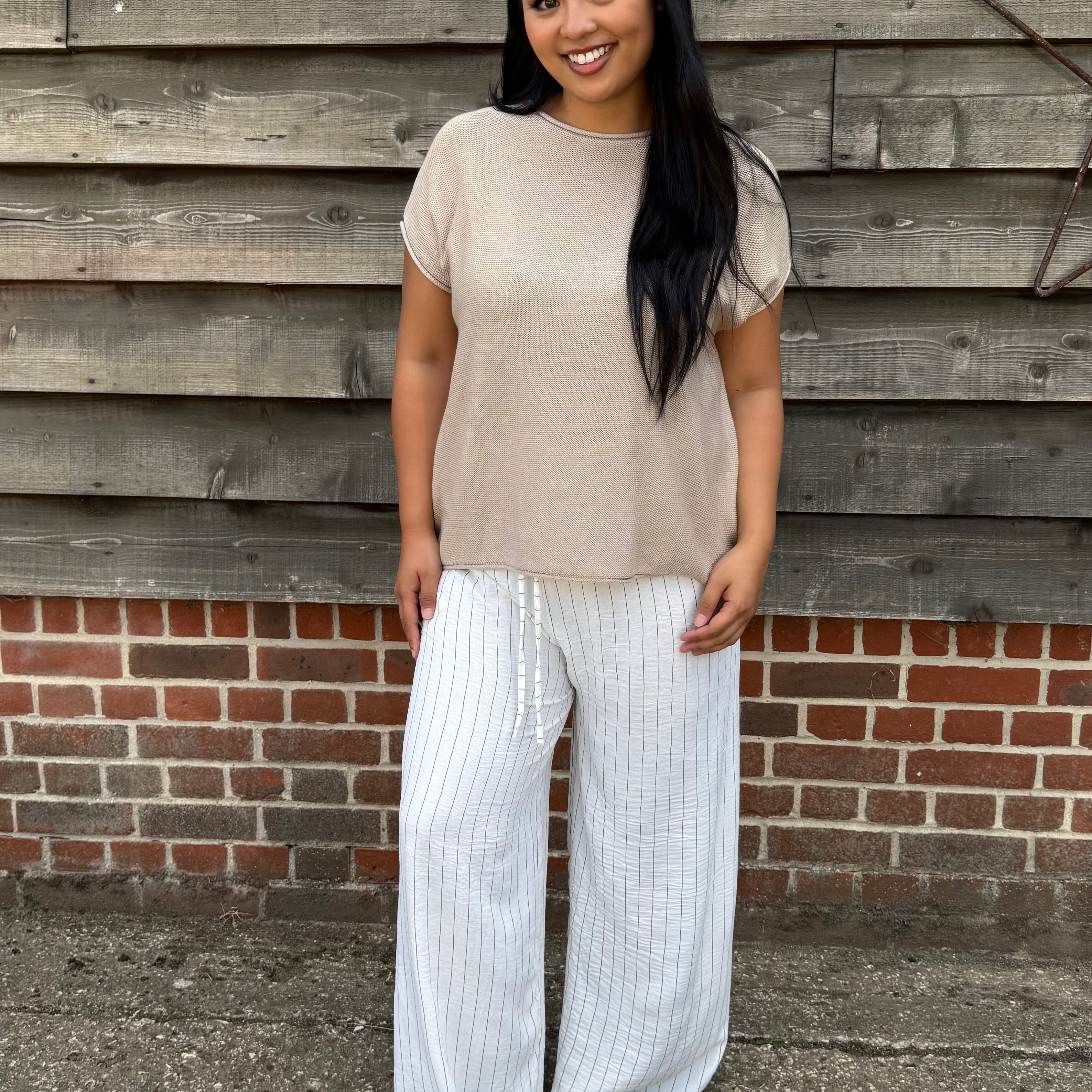 Woman wearing a beige top and white striped pants standing against a wooden wall.