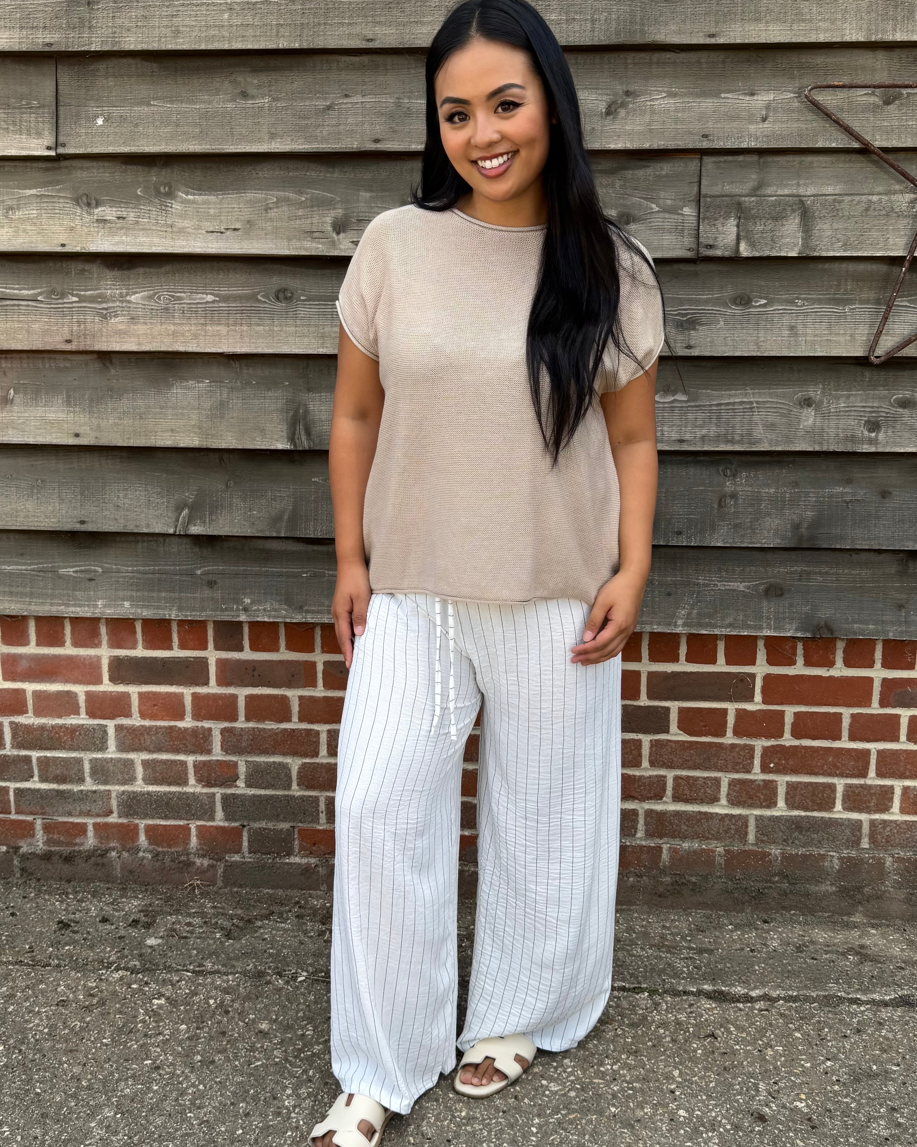 Woman wearing a beige top and white striped pants standing against a wooden wall.