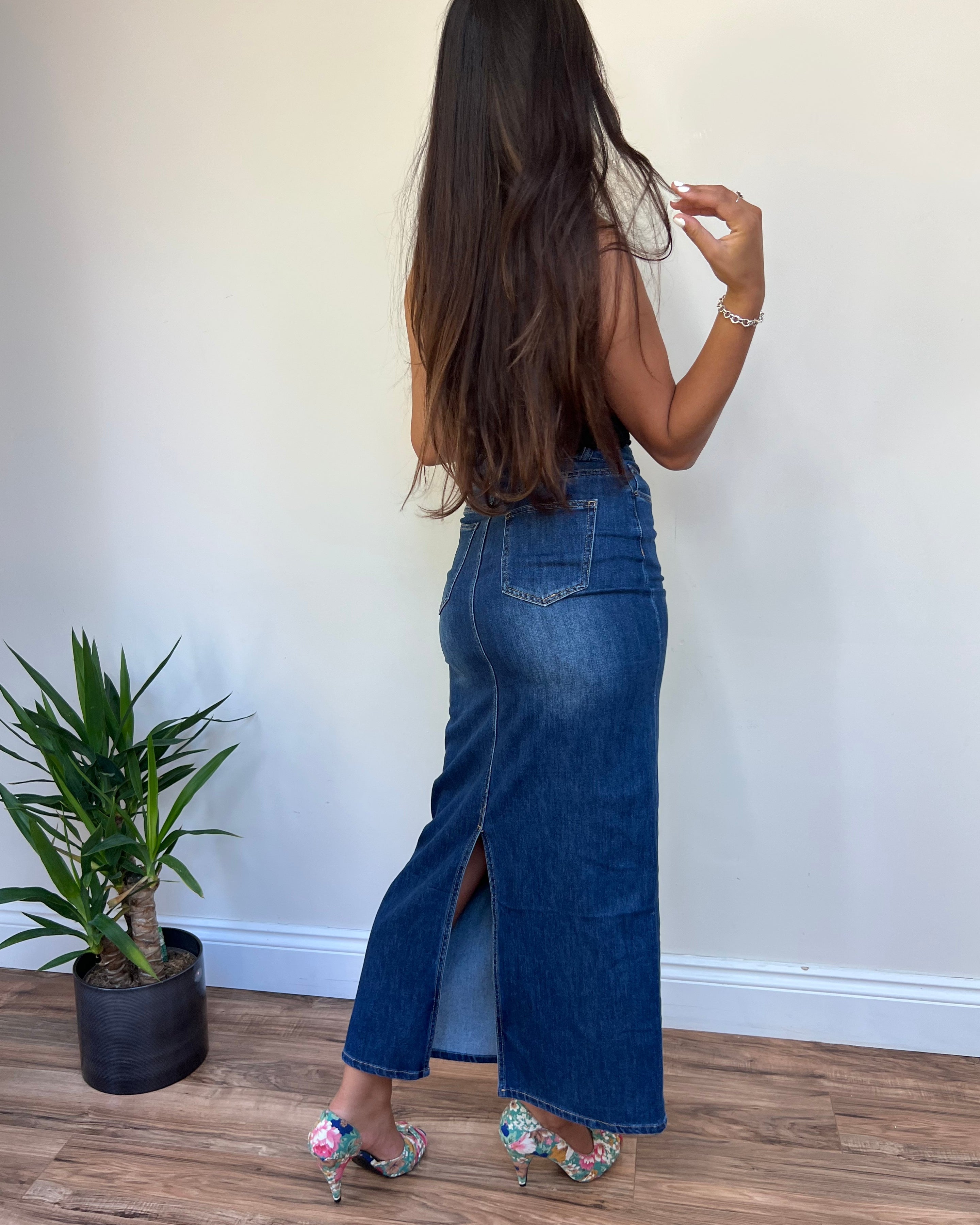A person standing with their back to the camera wearing a denim skirt with a split at the back, paired with floral shoes and posing near a potted plant.