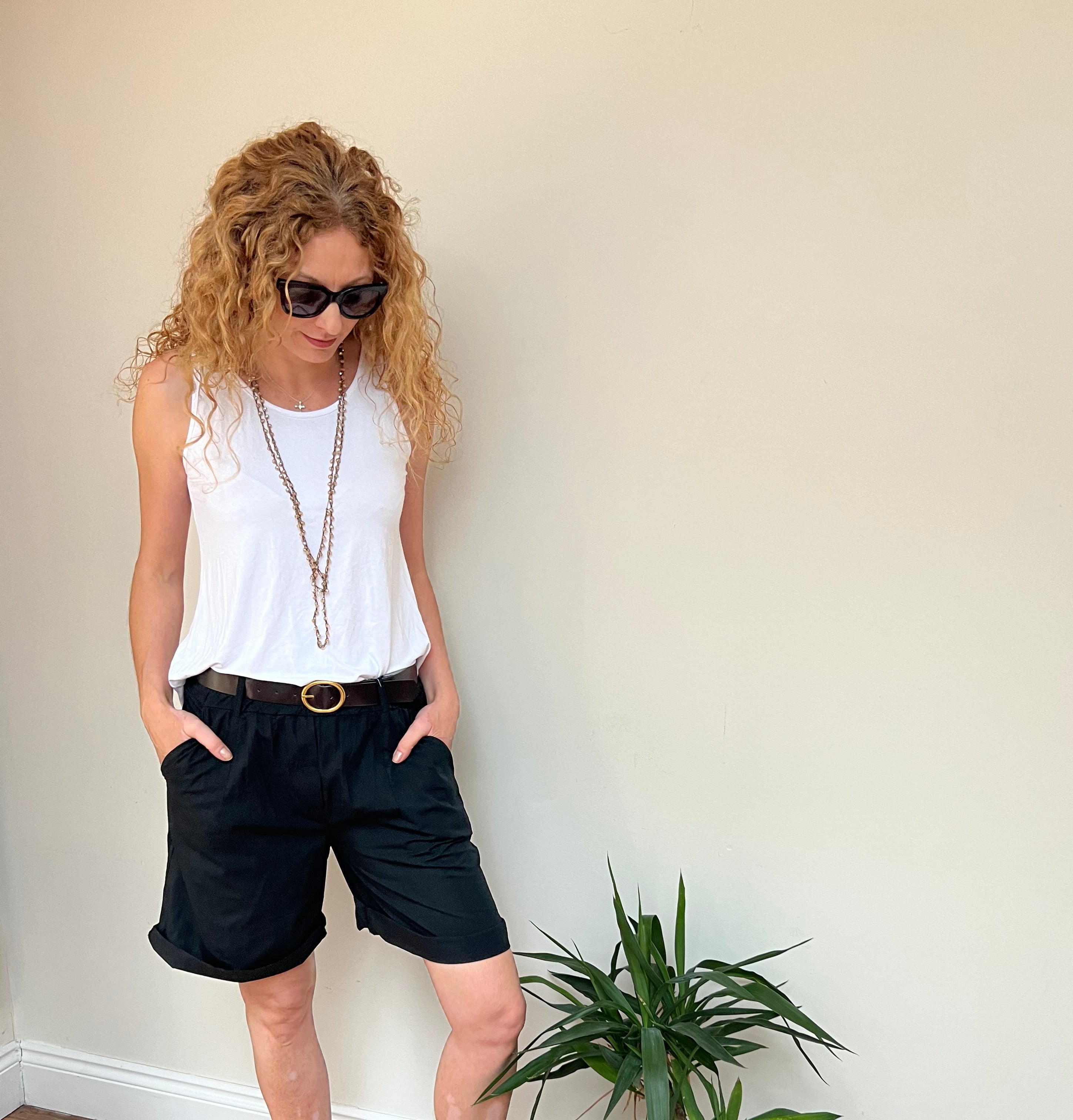 A person standing in a room with a white top and sunglasses, next to a potted plant, wearing black shorts with a brown belt.