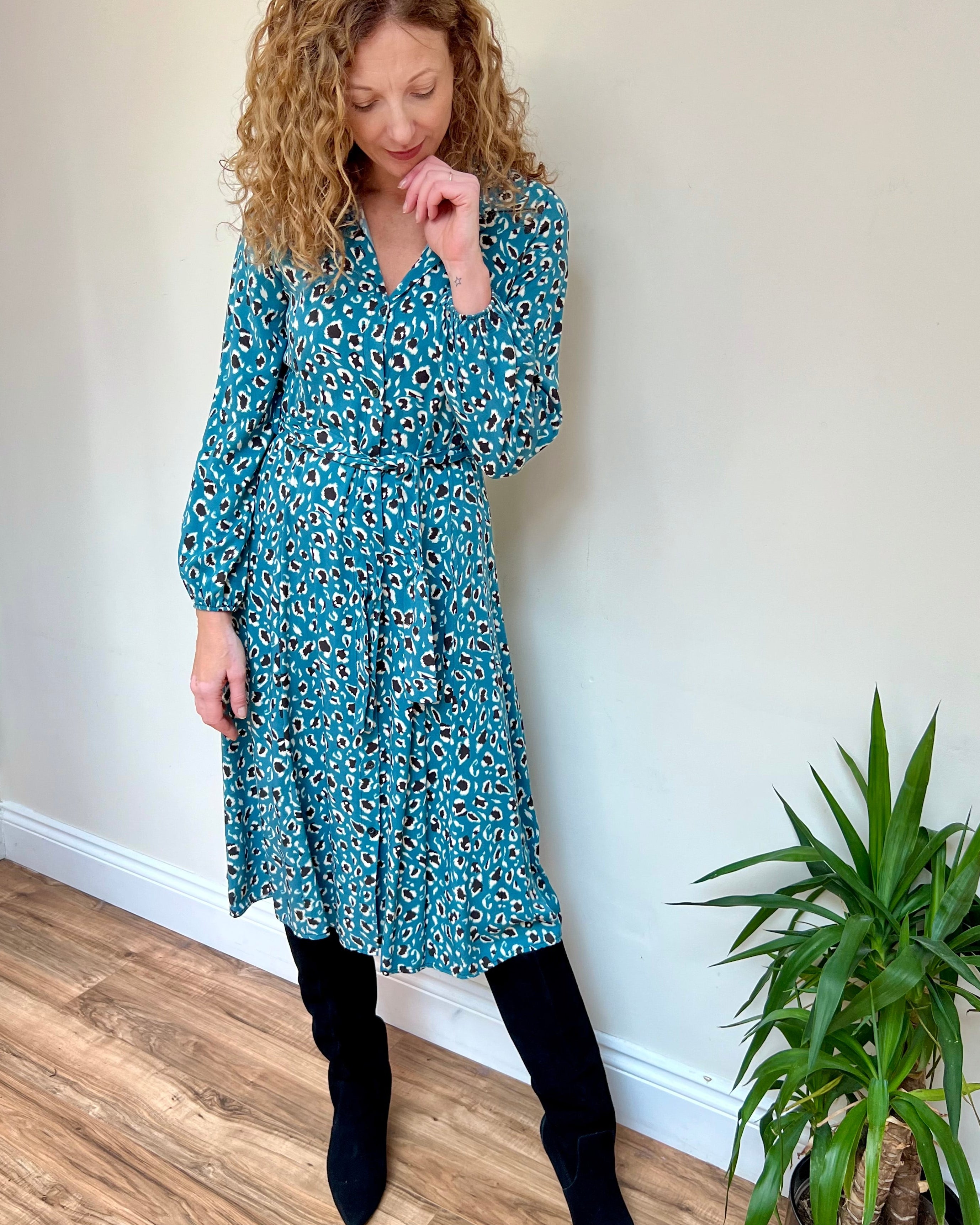 A woman wearing a midi teal animal print t-shirt dress with a tie waist, elasticated sleeves, collar, and a V-neckline, standing in a room with a potted plant beside her.