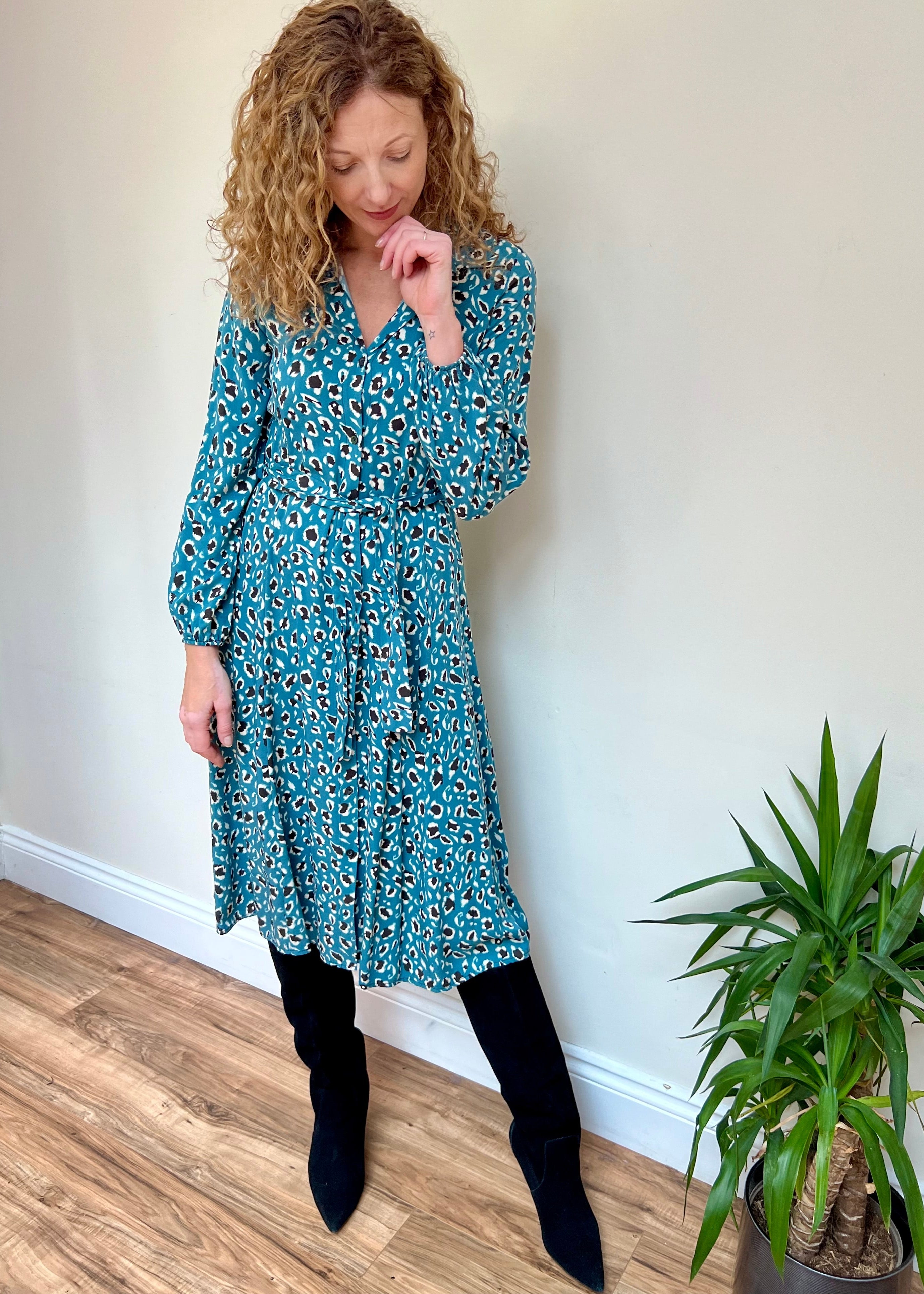 A woman wearing a midi teal animal print t-shirt dress with a tie waist, elasticated sleeves, collar, and a V-neckline, standing in a room with a potted plant beside her.