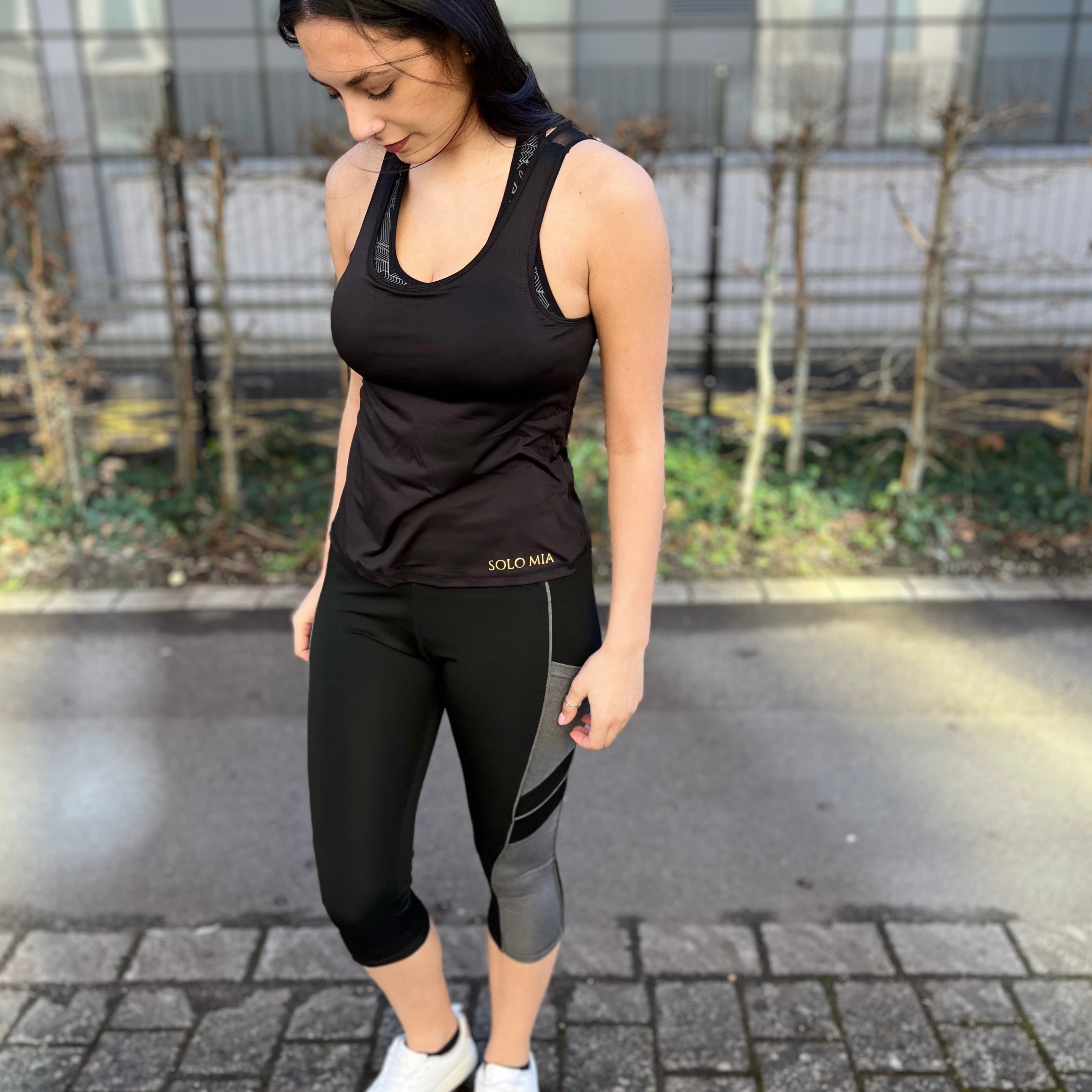 A person wearing black 3/4 length leggings with side pockets, paired with a sleeveless top and white sneakers, standing on a brick pathway.