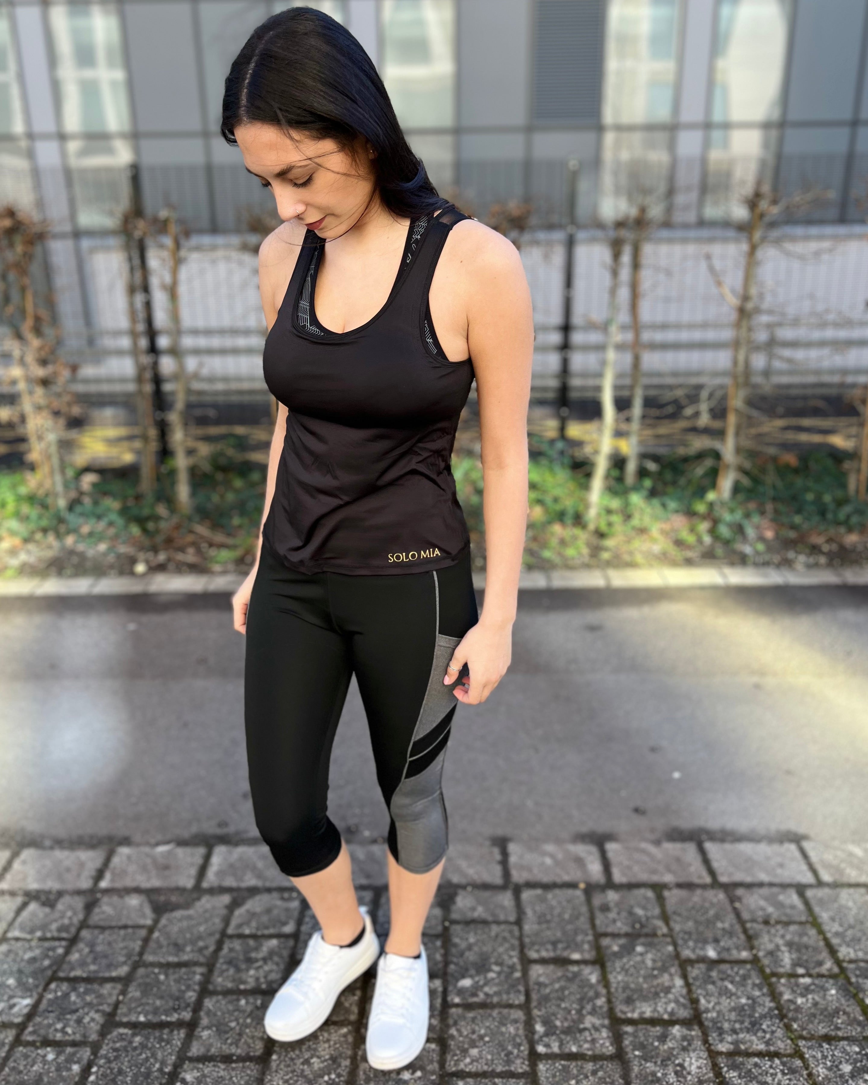 A person wearing black 3/4 length leggings with side pockets, paired with a sleeveless top and white sneakers, standing on a brick pathway.