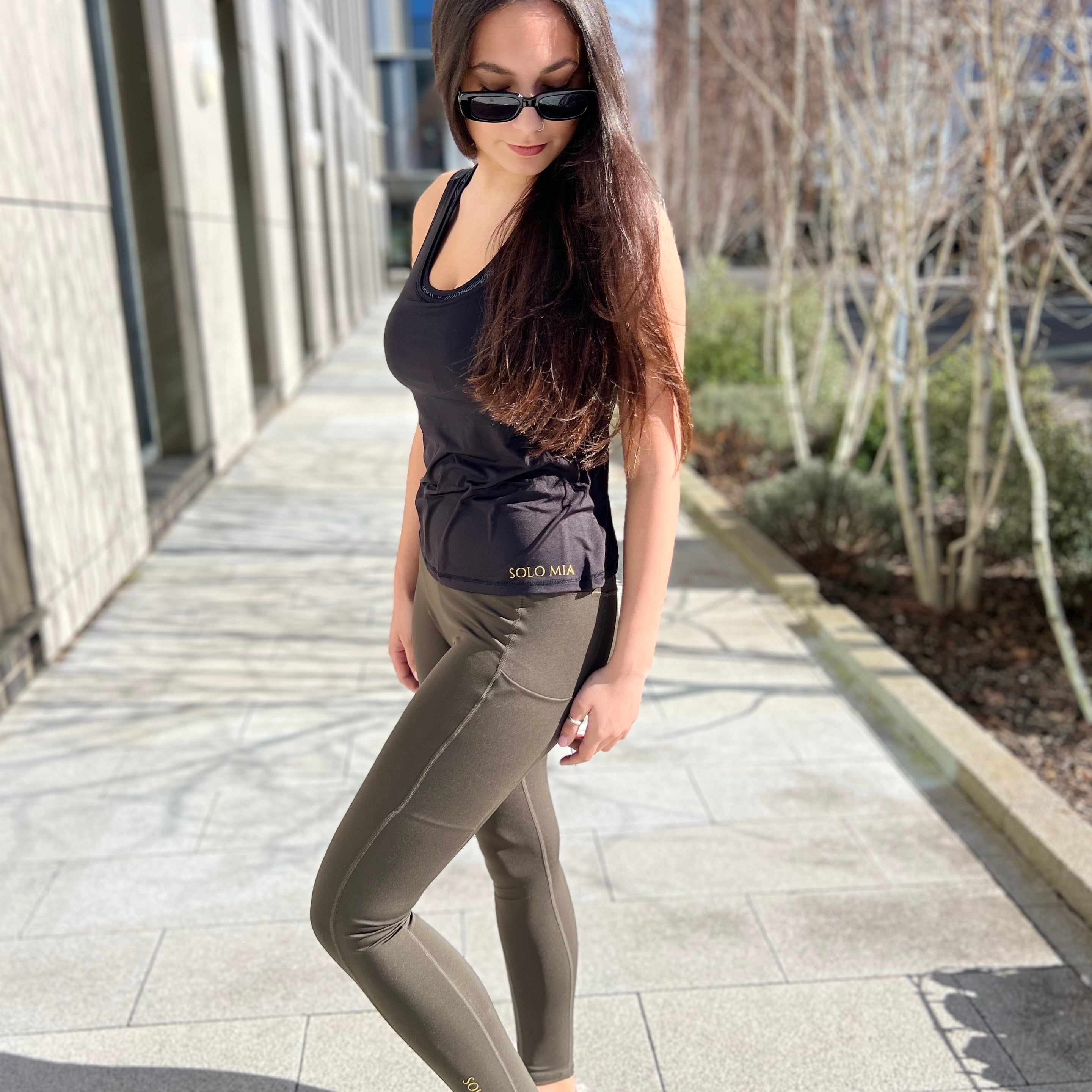 A woman standing on a sidewalk wearing sportiva running leggings, a tank top, sunglasses, and white sneakers.