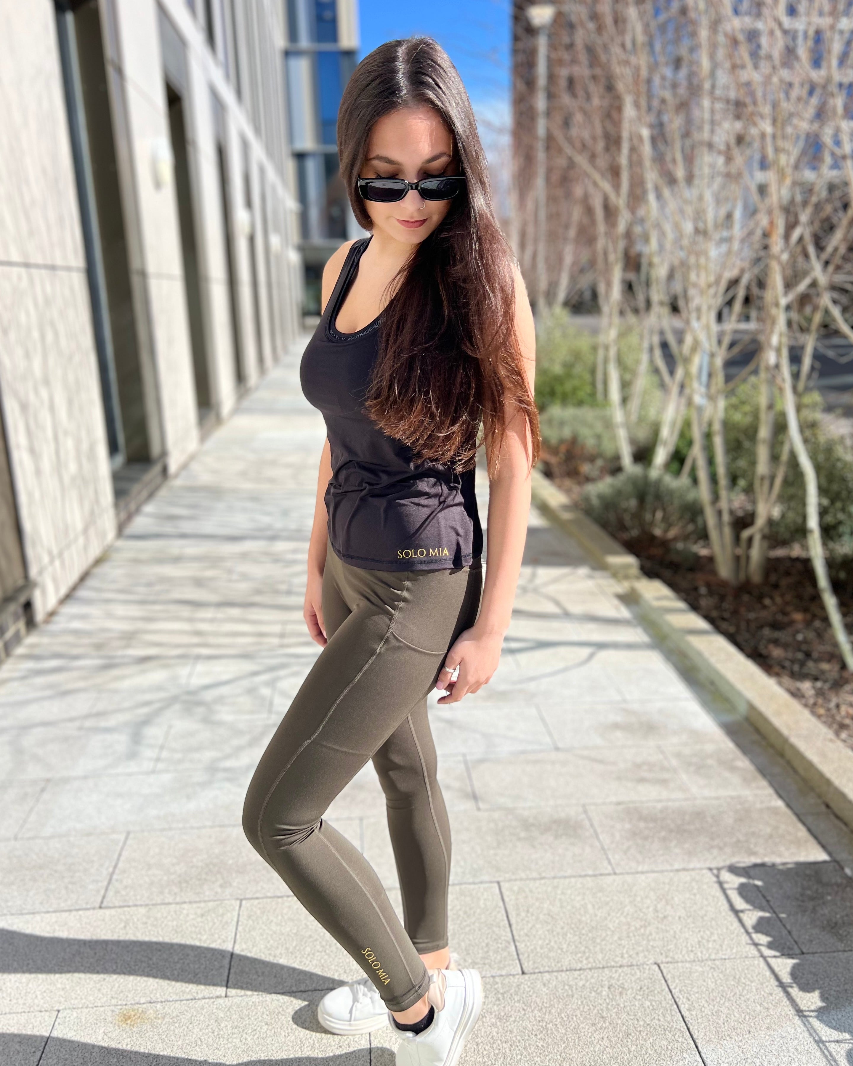A woman standing on a sidewalk wearing sportiva running leggings, a tank top, sunglasses, and white sneakers.