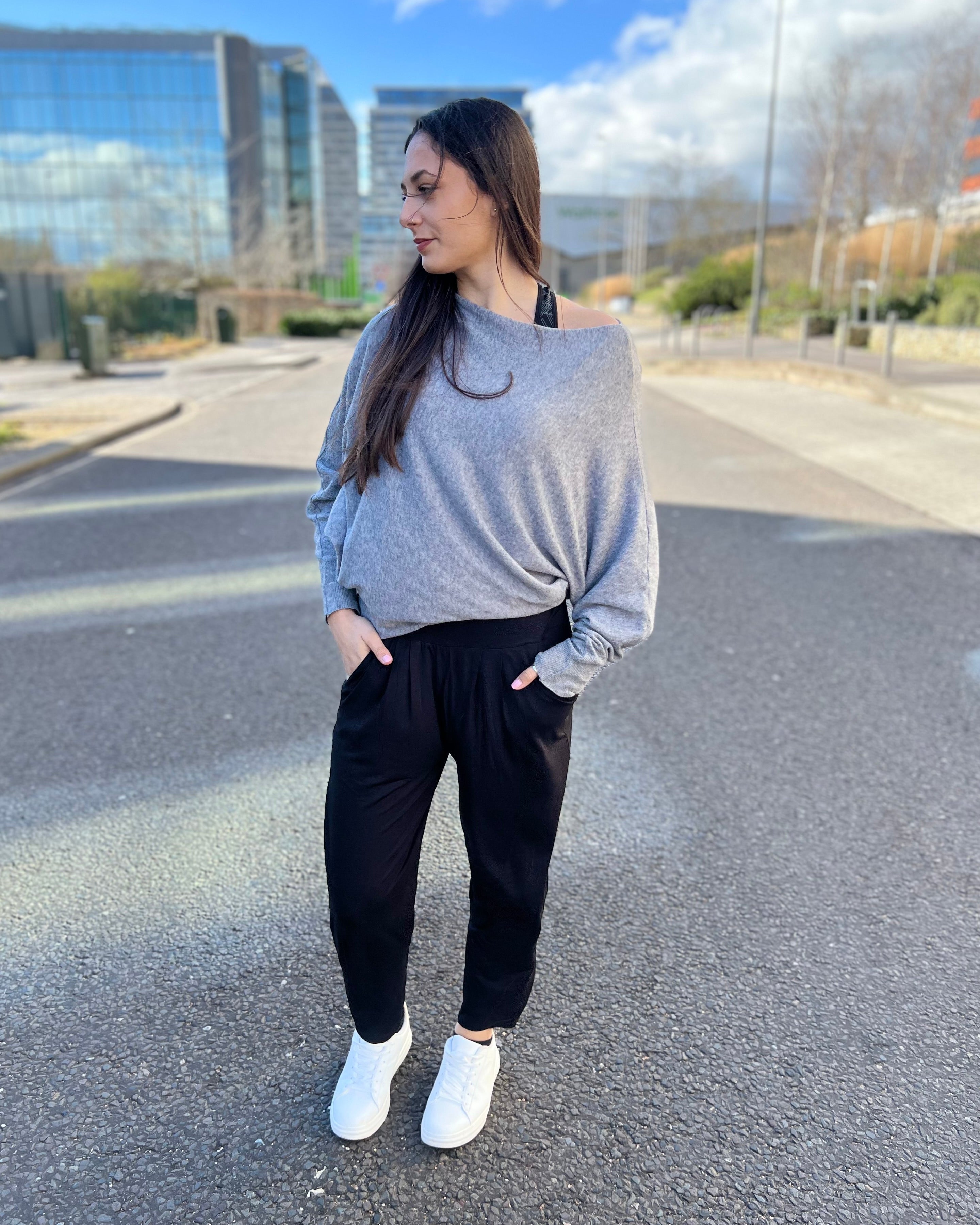 A woman standing on the street wearing black slouch joggers and white sneakers, with a grey sky and buildings in the background.