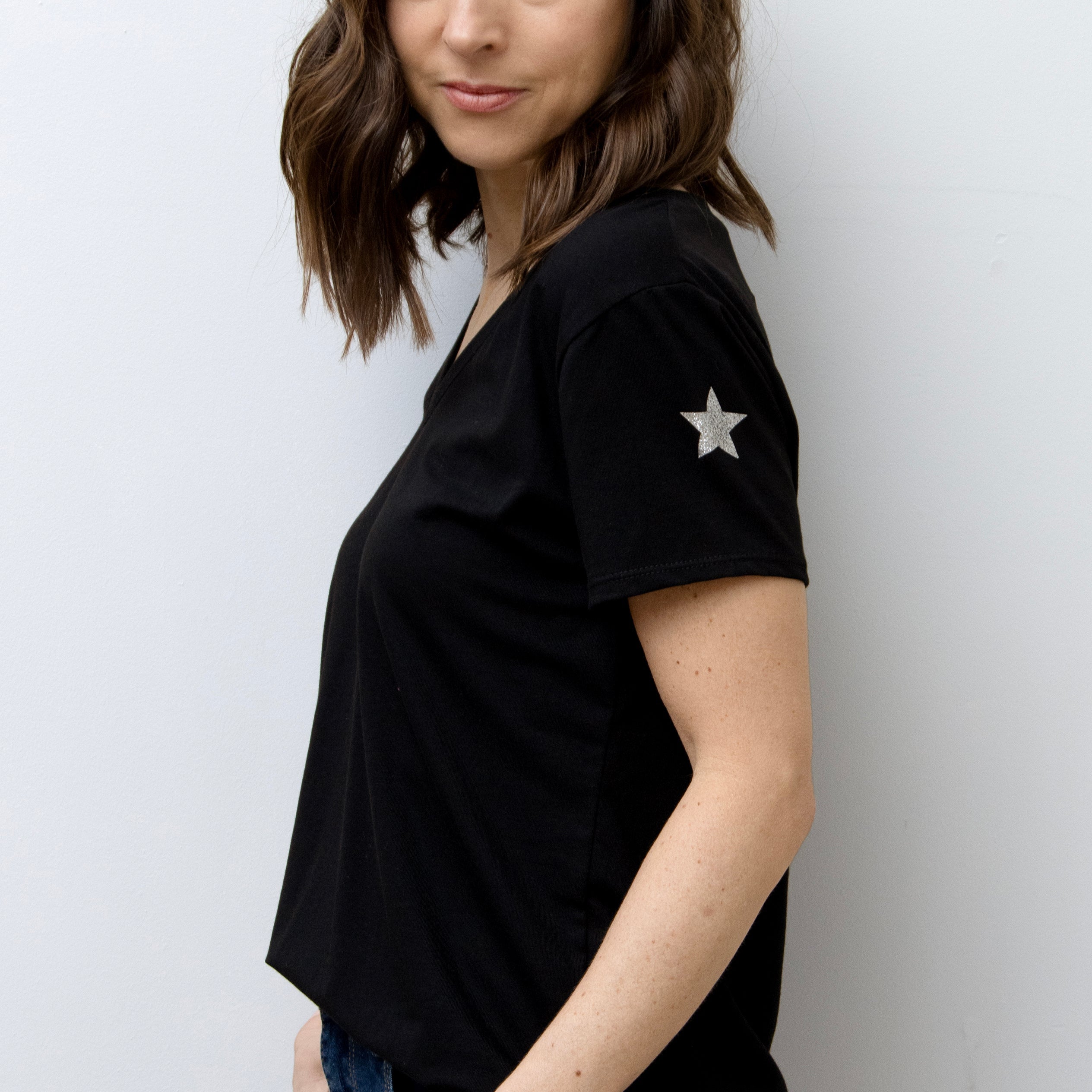 A woman smiling, wearing a pink V-neck T-shirt with a silver star on the sleeve, paired with blue jeans.