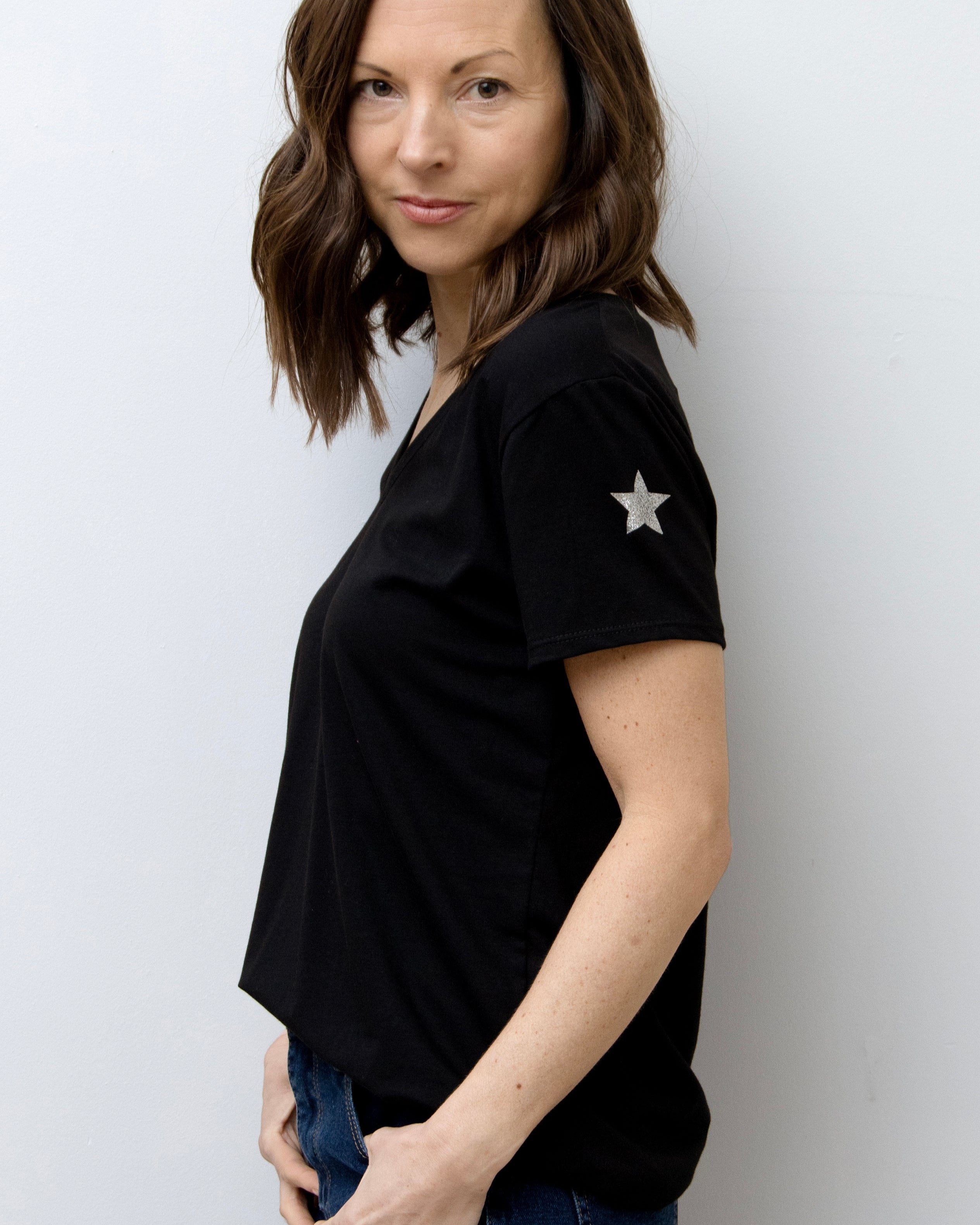 A woman smiling, wearing a pink V-neck T-shirt with a silver star on the sleeve, paired with blue jeans.