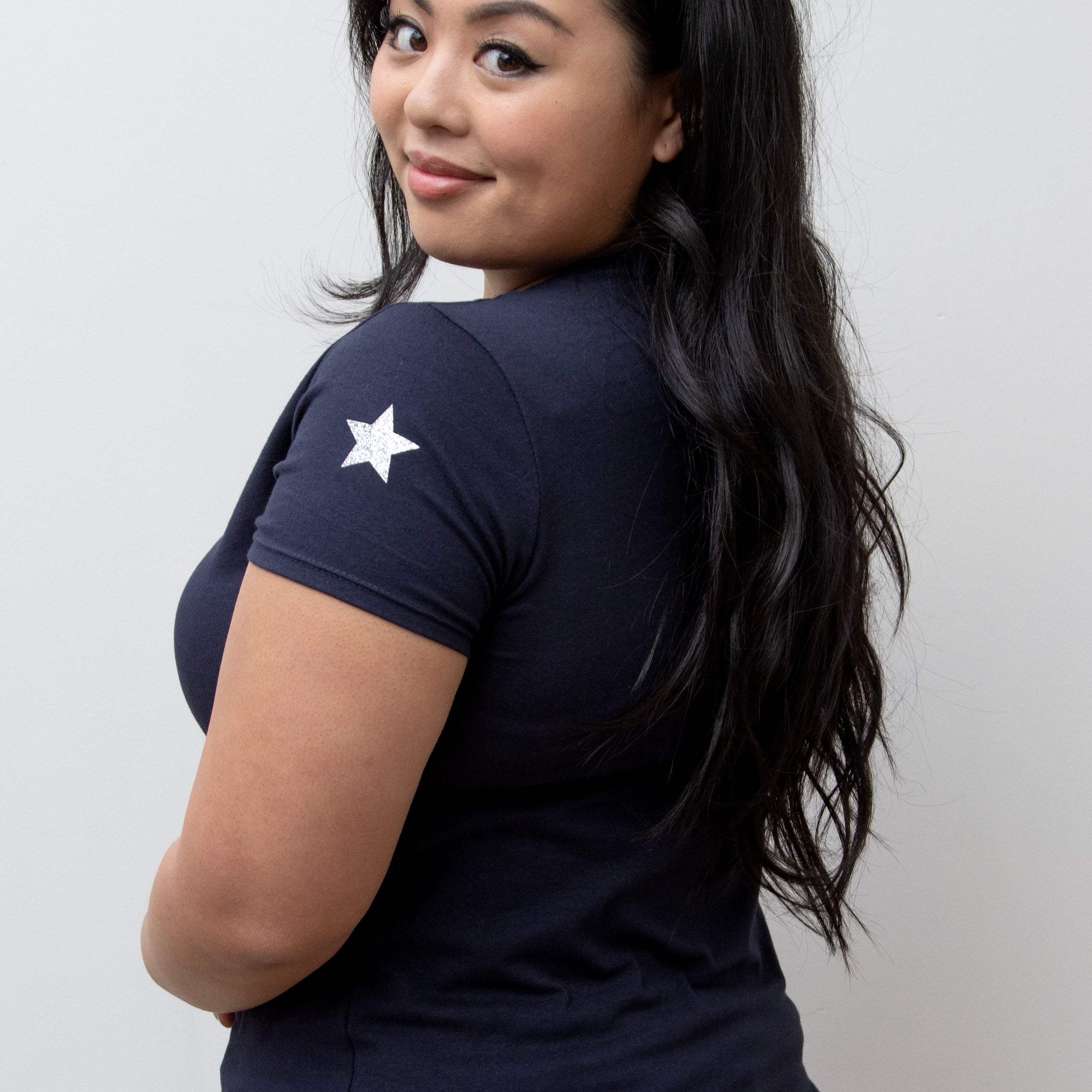A woman smiling, wearing a pink V-neck T-shirt with a silver star on the sleeve, paired with blue jeans.