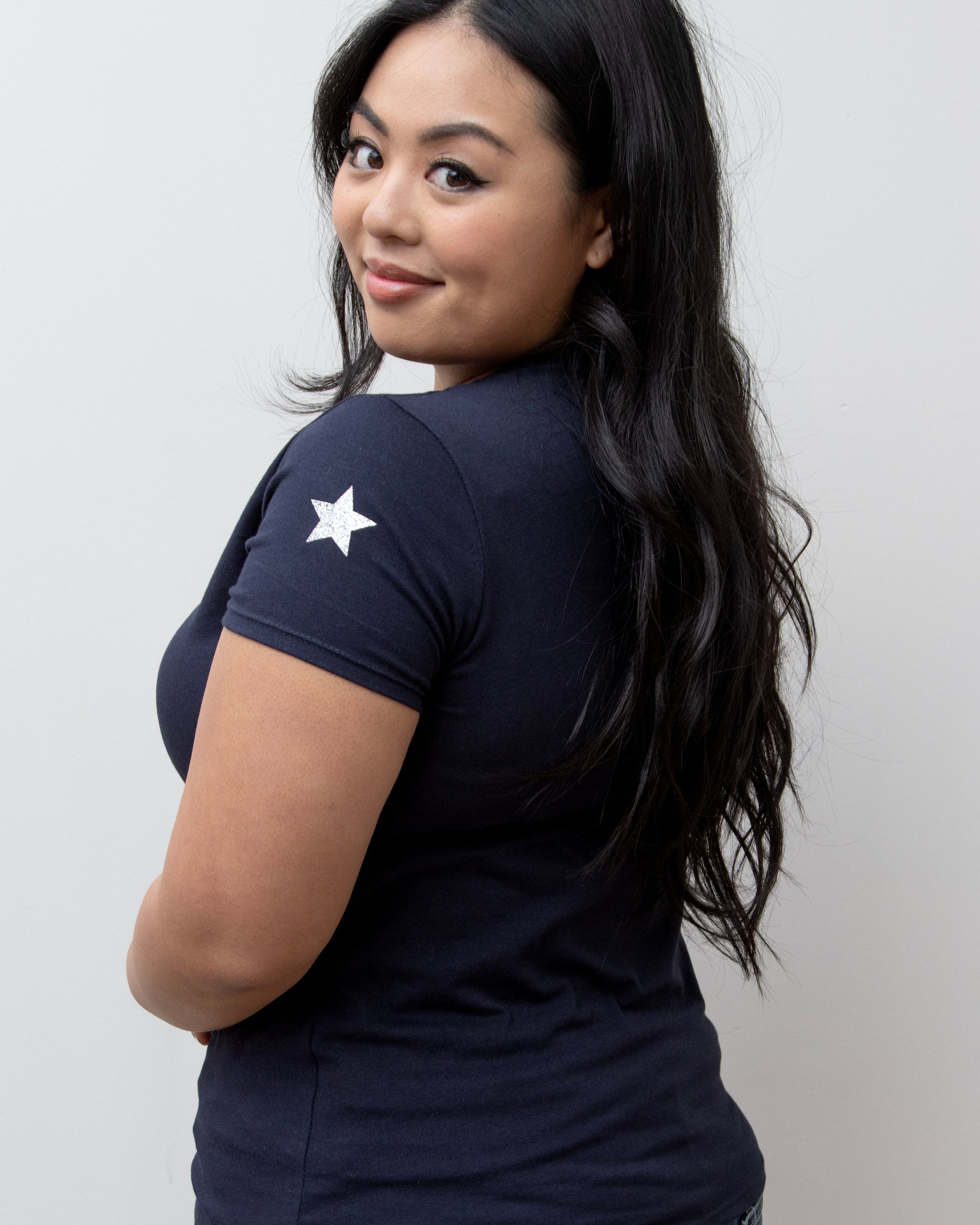 A woman smiling, wearing a pink V-neck T-shirt with a silver star on the sleeve, paired with blue jeans.
