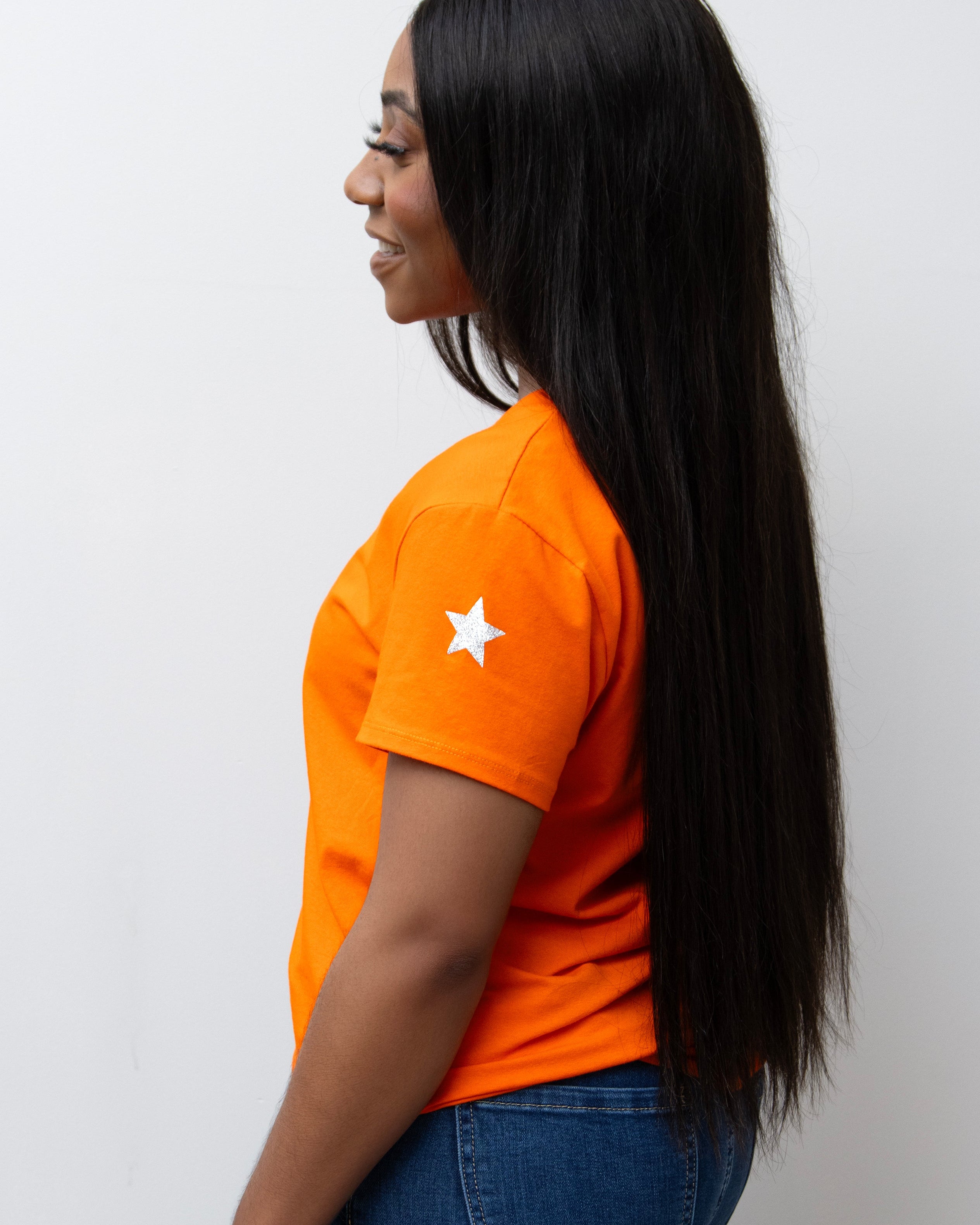A woman smiling, wearing a pink V-neck T-shirt with a silver star on the sleeve, paired with blue jeans.