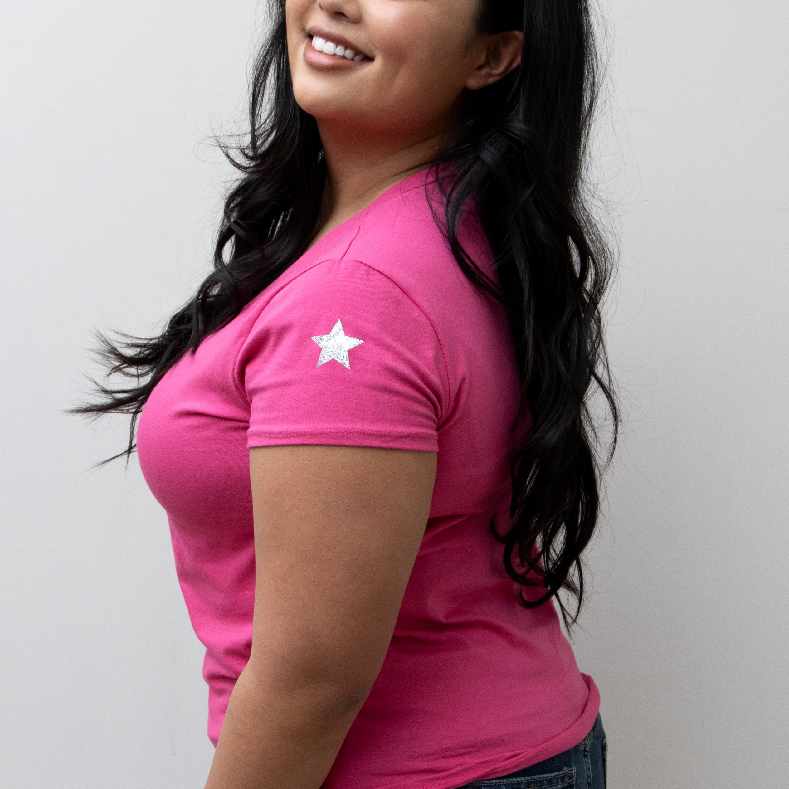 A woman smiling, wearing a pink V-neck T-shirt with a silver star on the sleeve, paired with blue jeans.