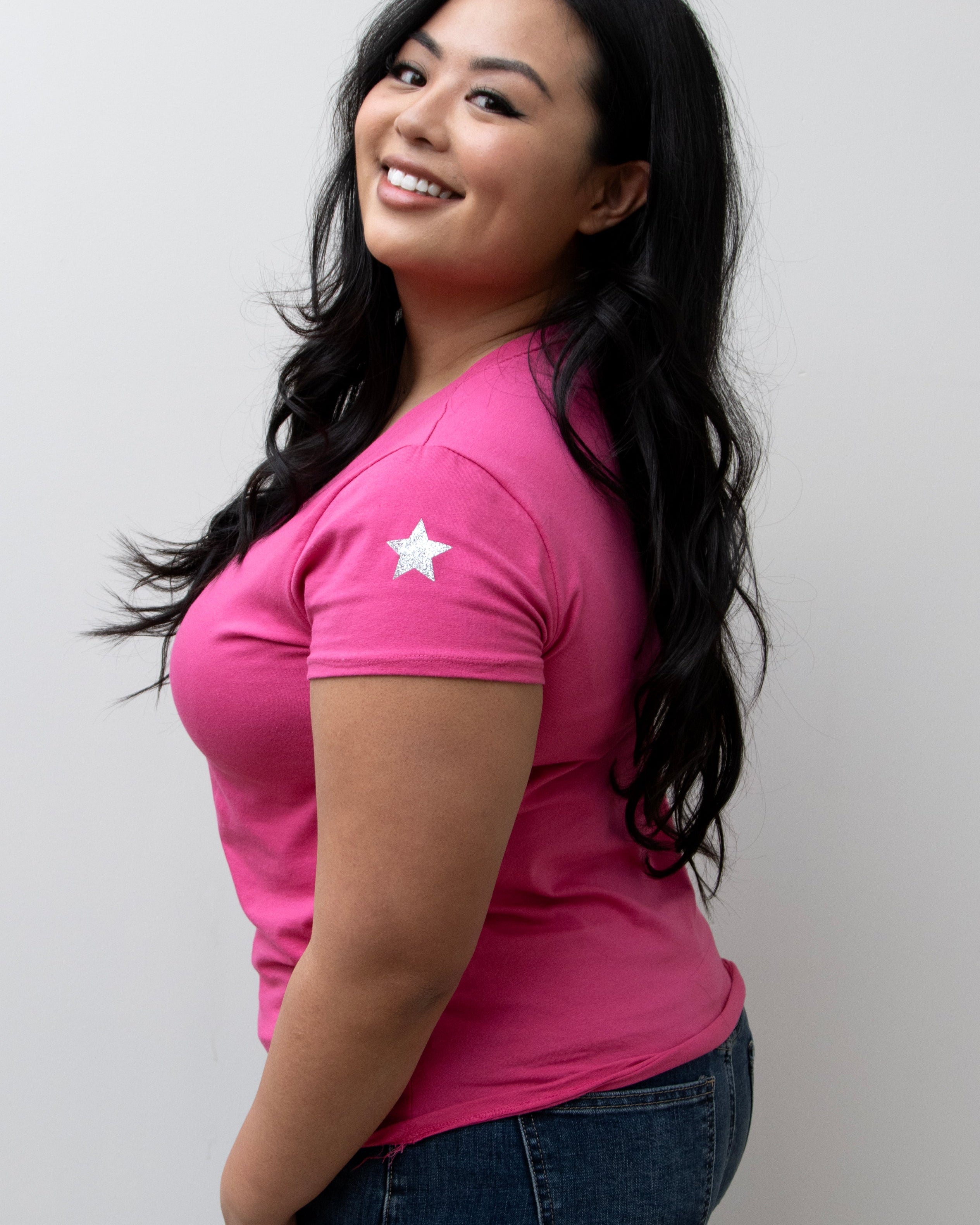 A woman smiling, wearing a pink V-neck T-shirt with a silver star on the sleeve, paired with blue jeans.