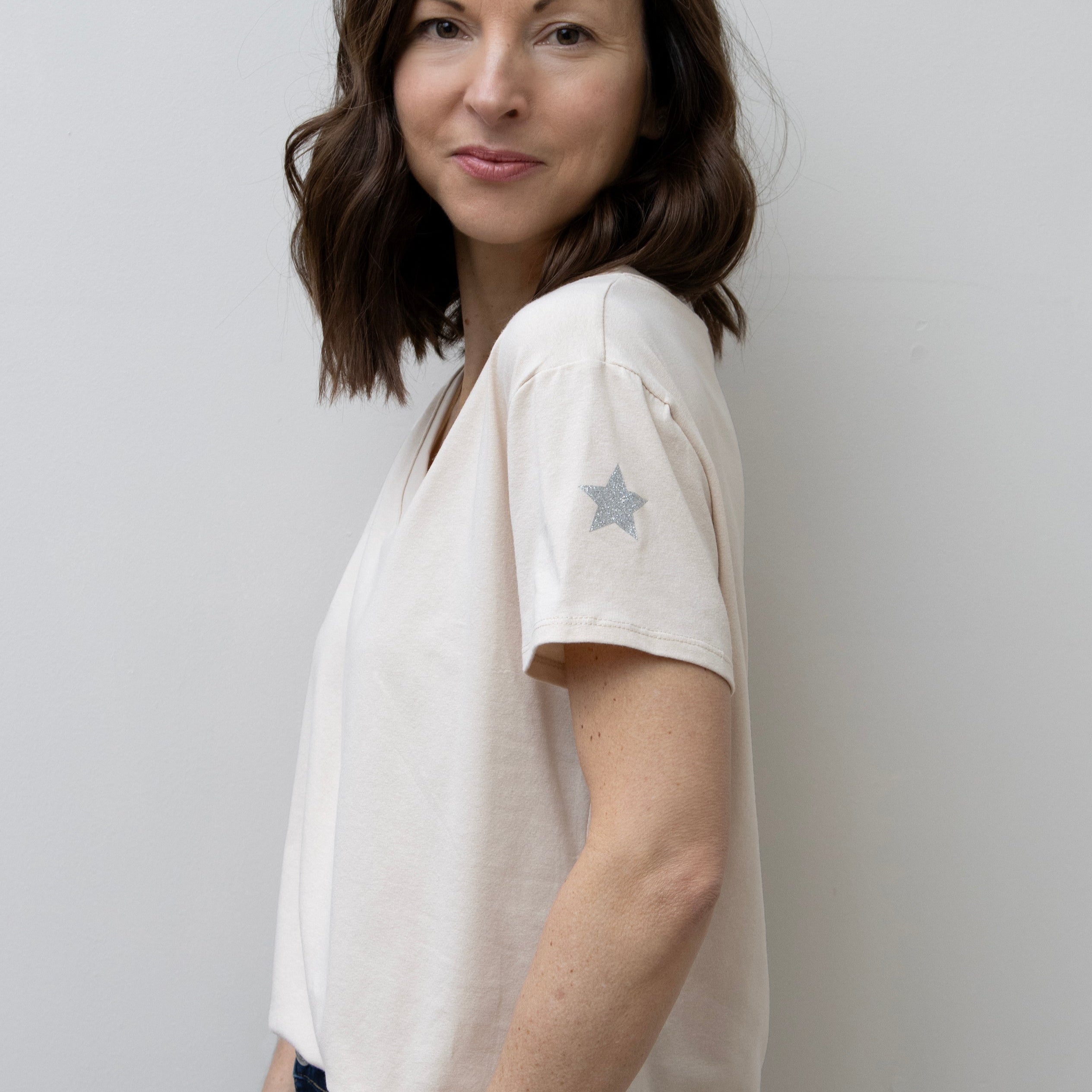 A woman smiling, wearing a pink V-neck T-shirt with a silver star on the sleeve, paired with blue jeans.
