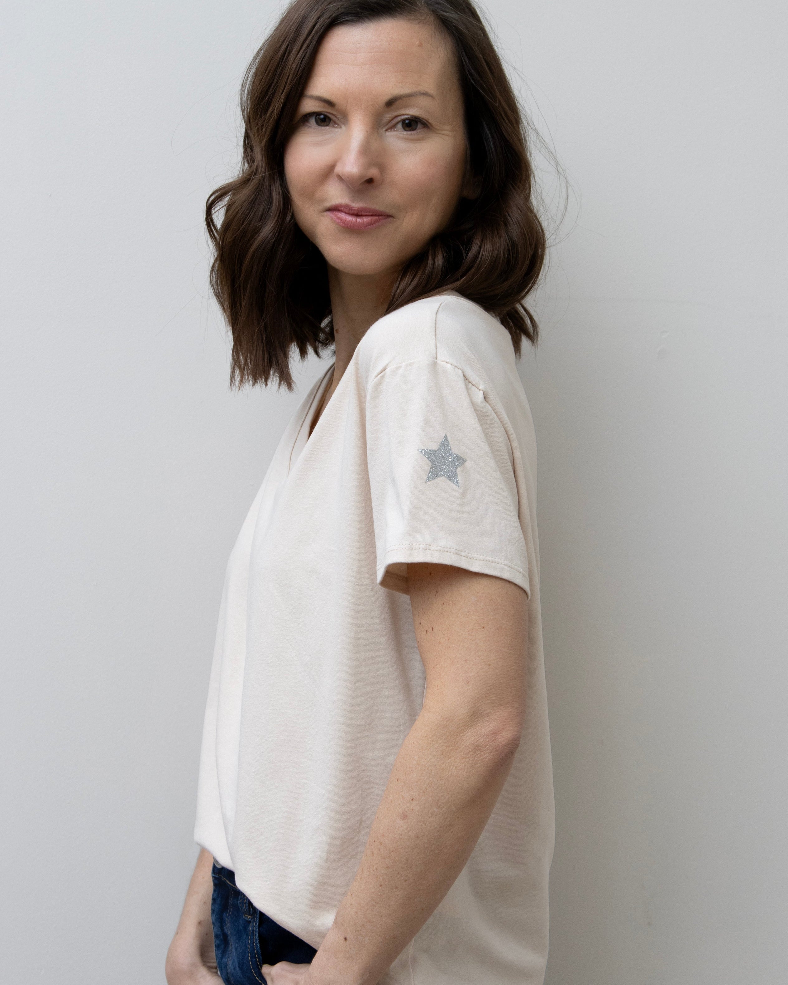 A woman smiling, wearing a pink V-neck T-shirt with a silver star on the sleeve, paired with blue jeans.
