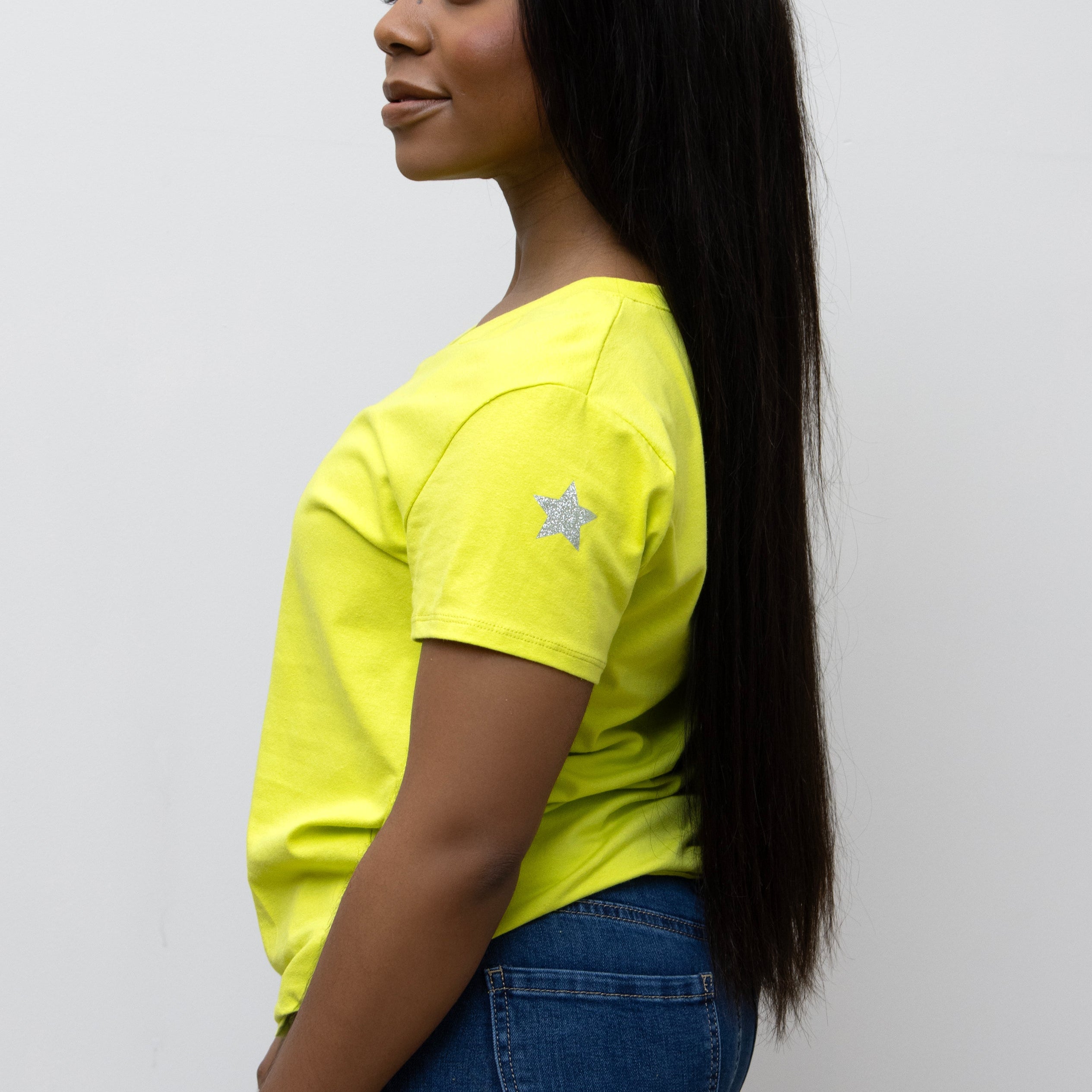 A woman smiling, wearing a pink V-neck T-shirt with a silver star on the sleeve, paired with blue jeans.