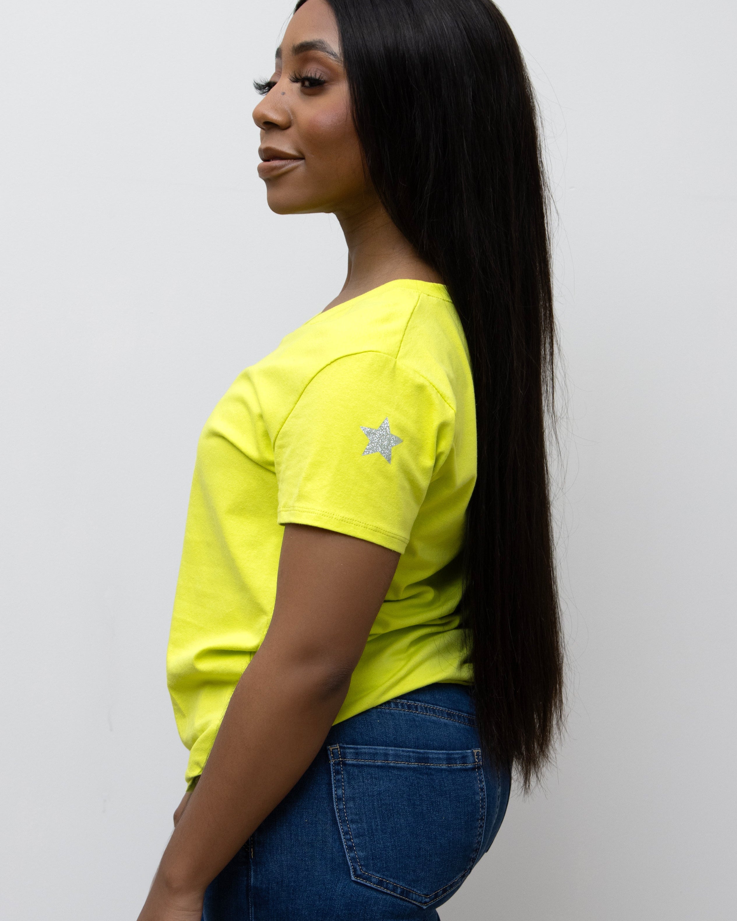 A woman smiling, wearing a pink V-neck T-shirt with a silver star on the sleeve, paired with blue jeans.