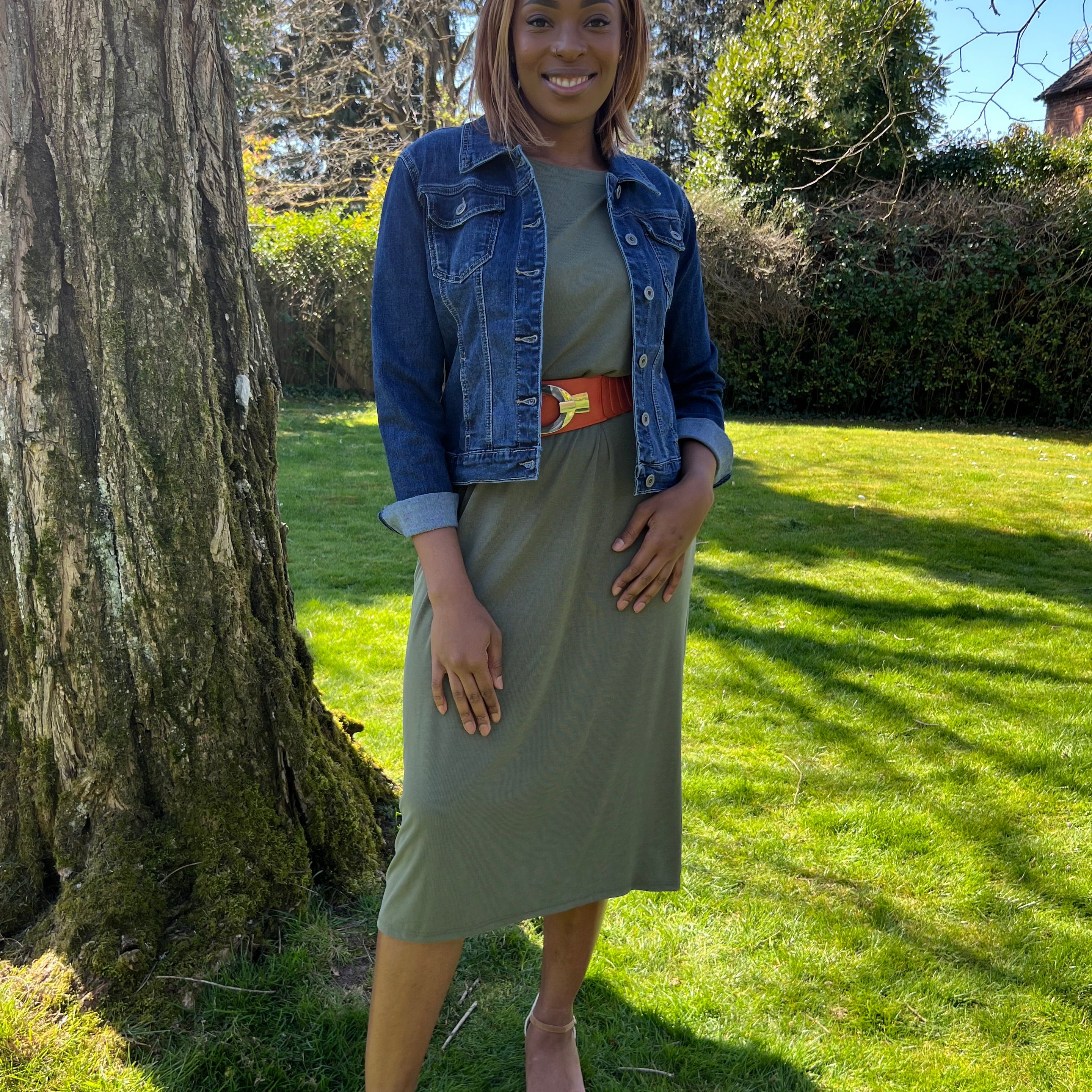 A woman standing outdoors wearing a green midi dress with a round neckline, sleeveless design, and knee-length. She has on a blue denim jacket, beige belt, and matching beige shoes.