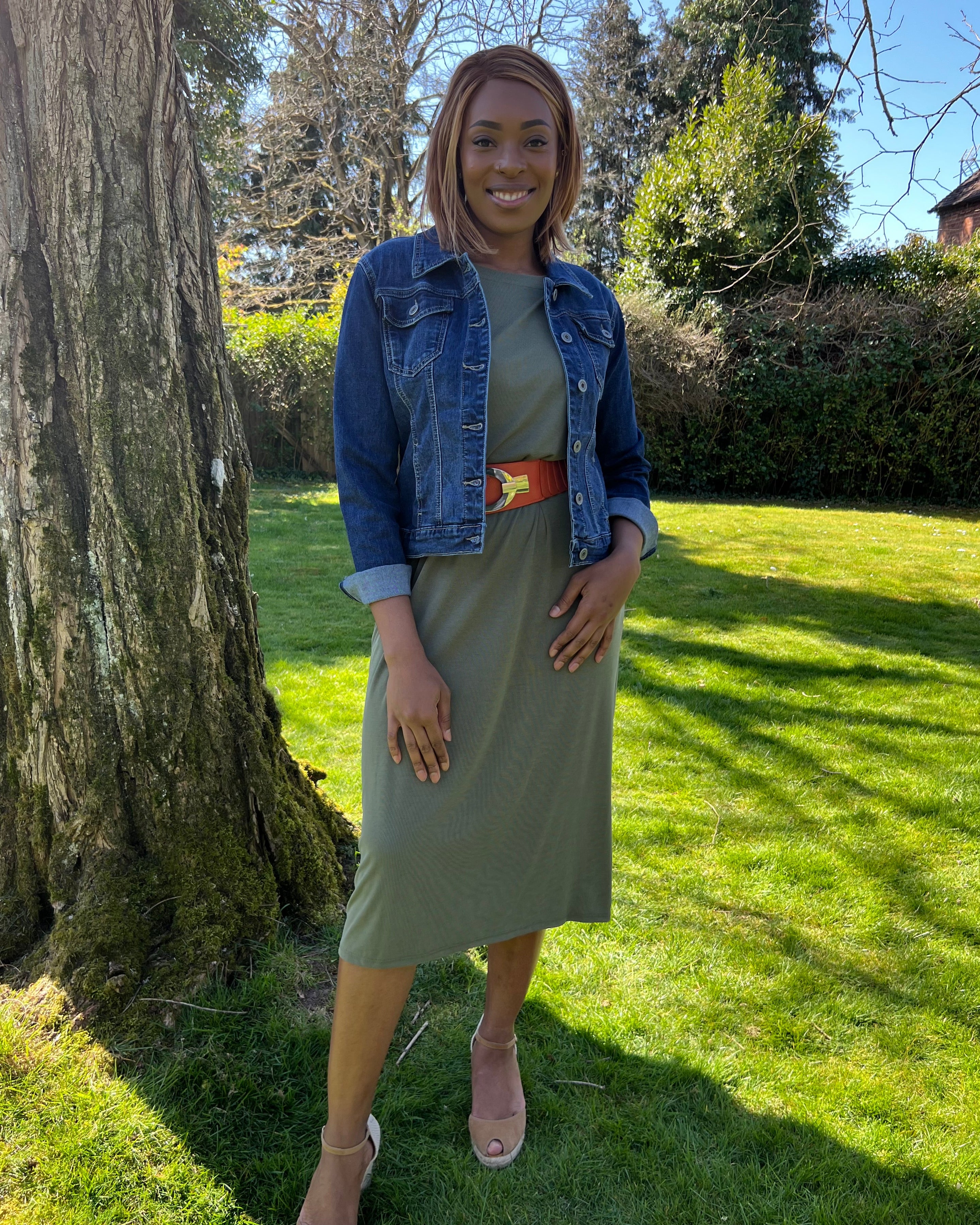 A woman standing outdoors wearing a green midi dress with a round neckline, sleeveless design, and knee-length. She has on a blue denim jacket, beige belt, and matching beige shoes.