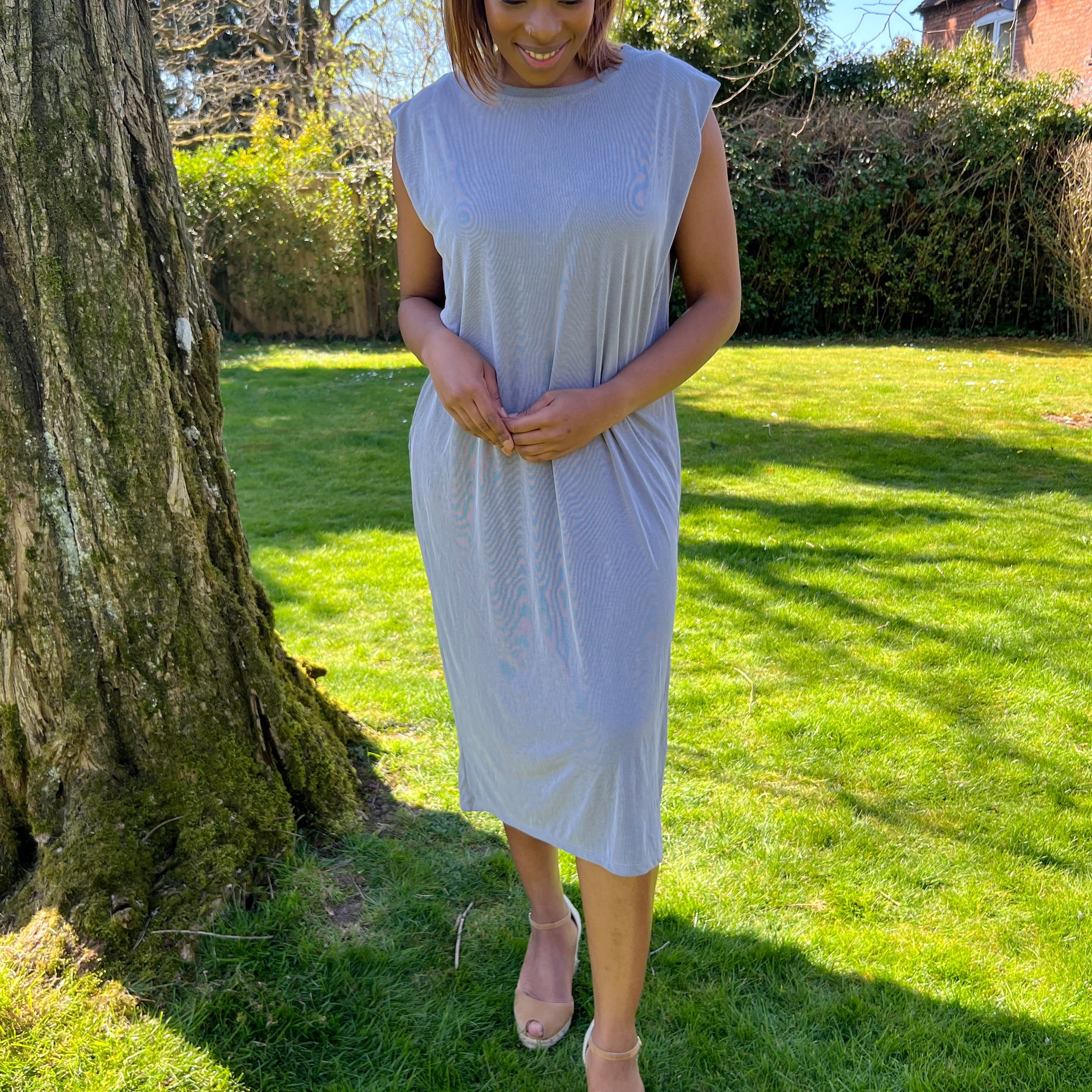 A woman standing outdoors wearing a green midi dress with a round neckline, sleeveless design, and knee-length. She has on a blue denim jacket, beige belt, and matching beige shoes.