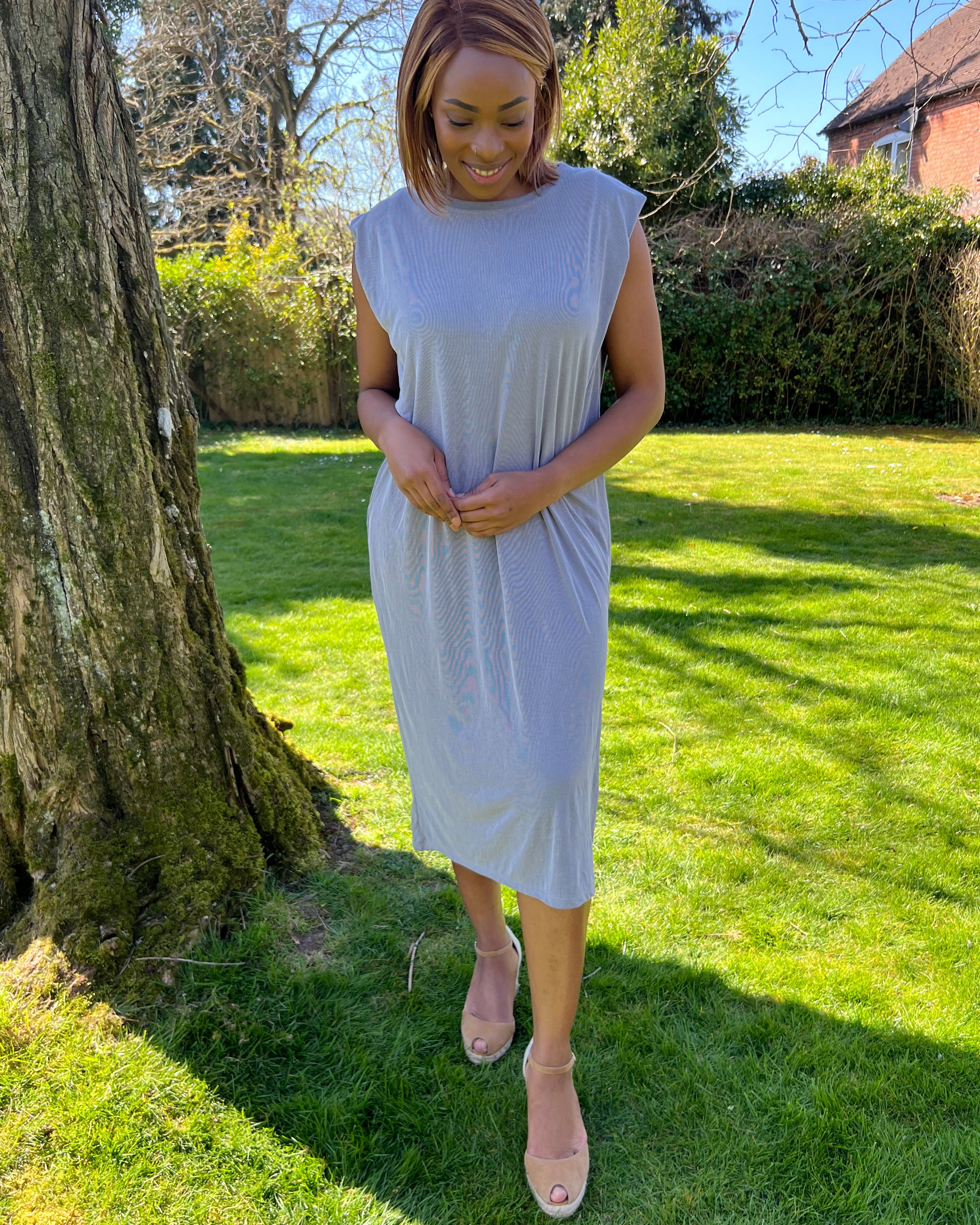 A woman standing outdoors wearing a green midi dress with a round neckline, sleeveless design, and knee-length. She has on a blue denim jacket, beige belt, and matching beige shoes.