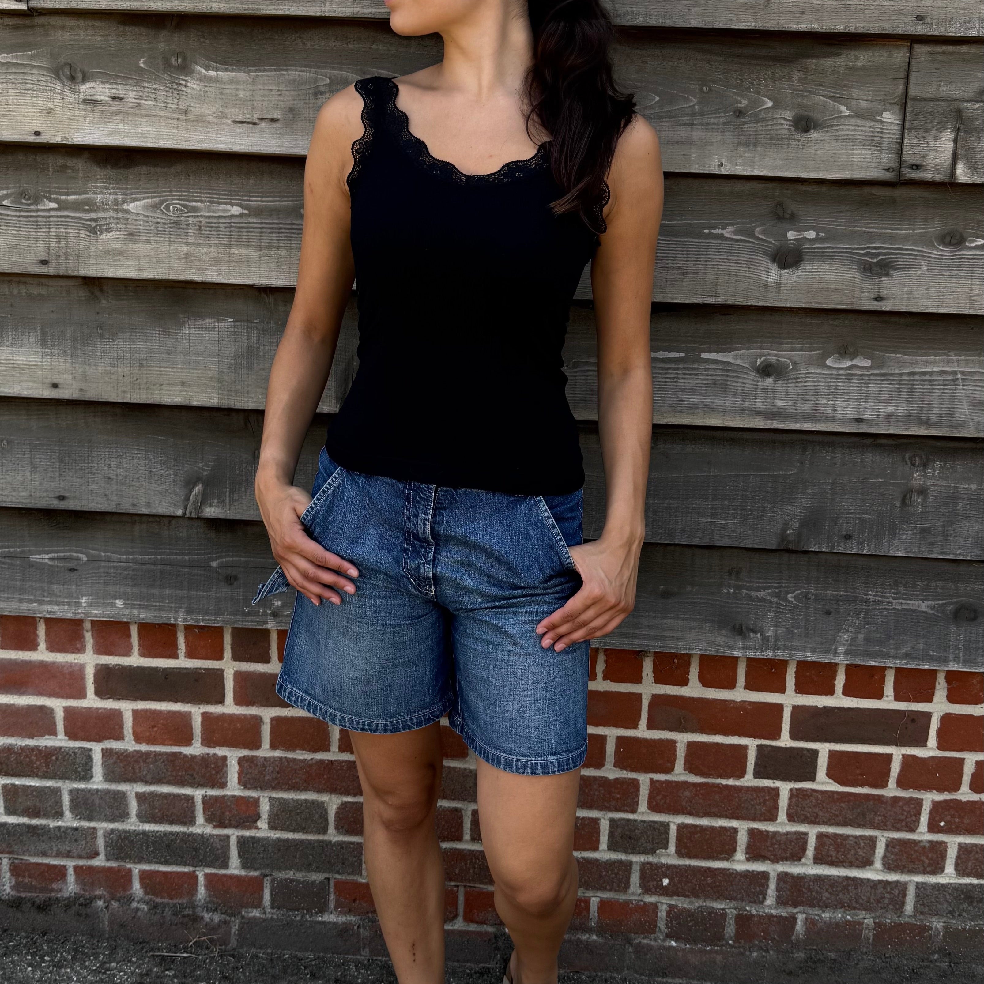 Woman wearing a black top and denim shorts standing against a wooden wall.