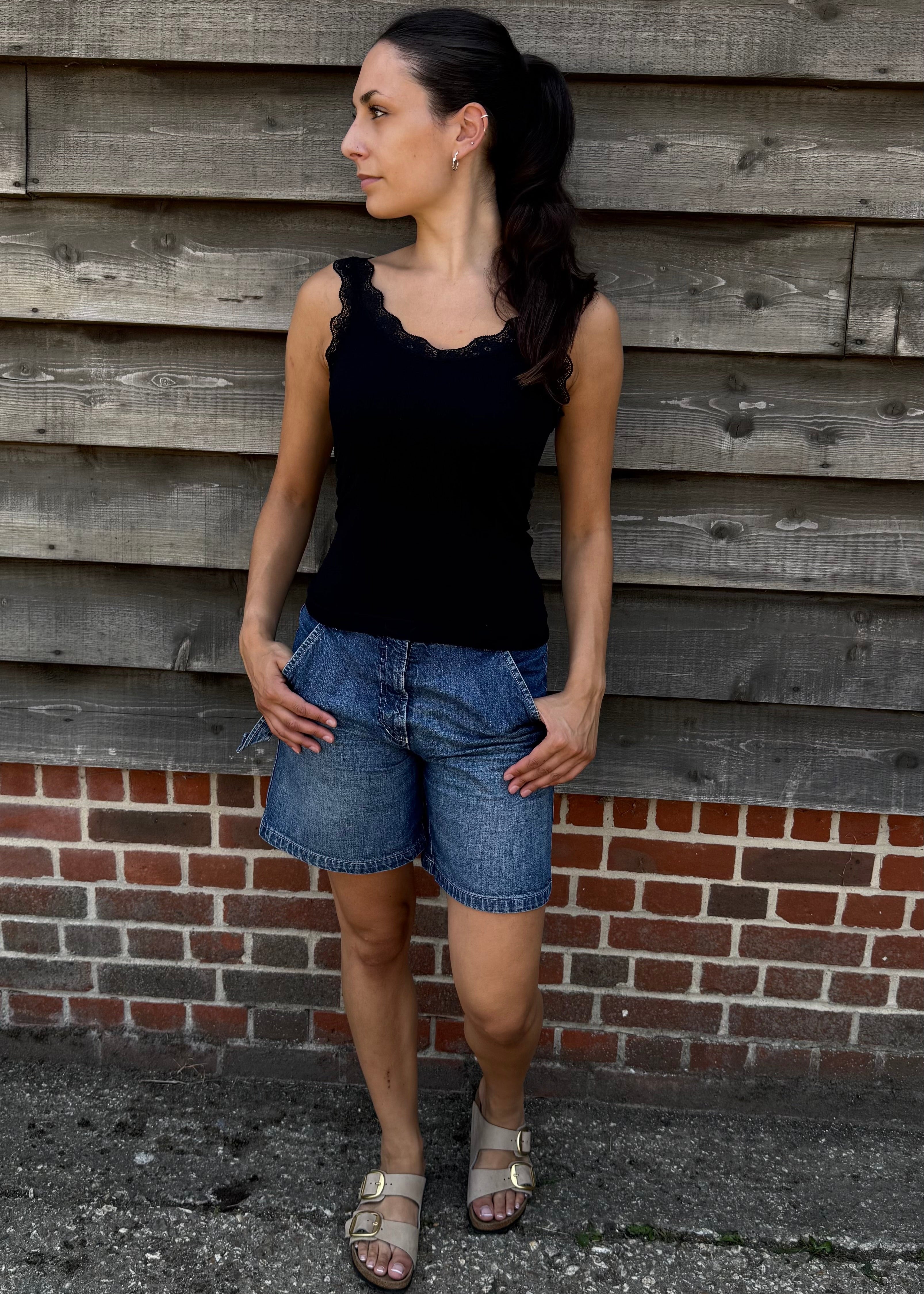 Woman wearing a black top and denim shorts standing against a wooden wall.