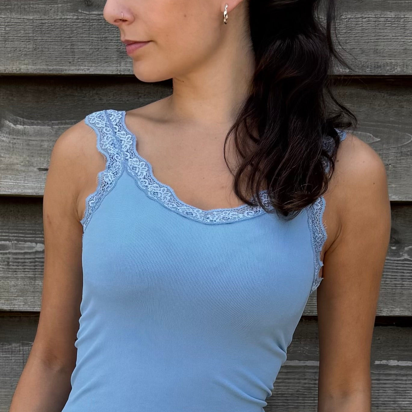 Woman wearing a blue tank top and denim shorts standing against a wooden wall.