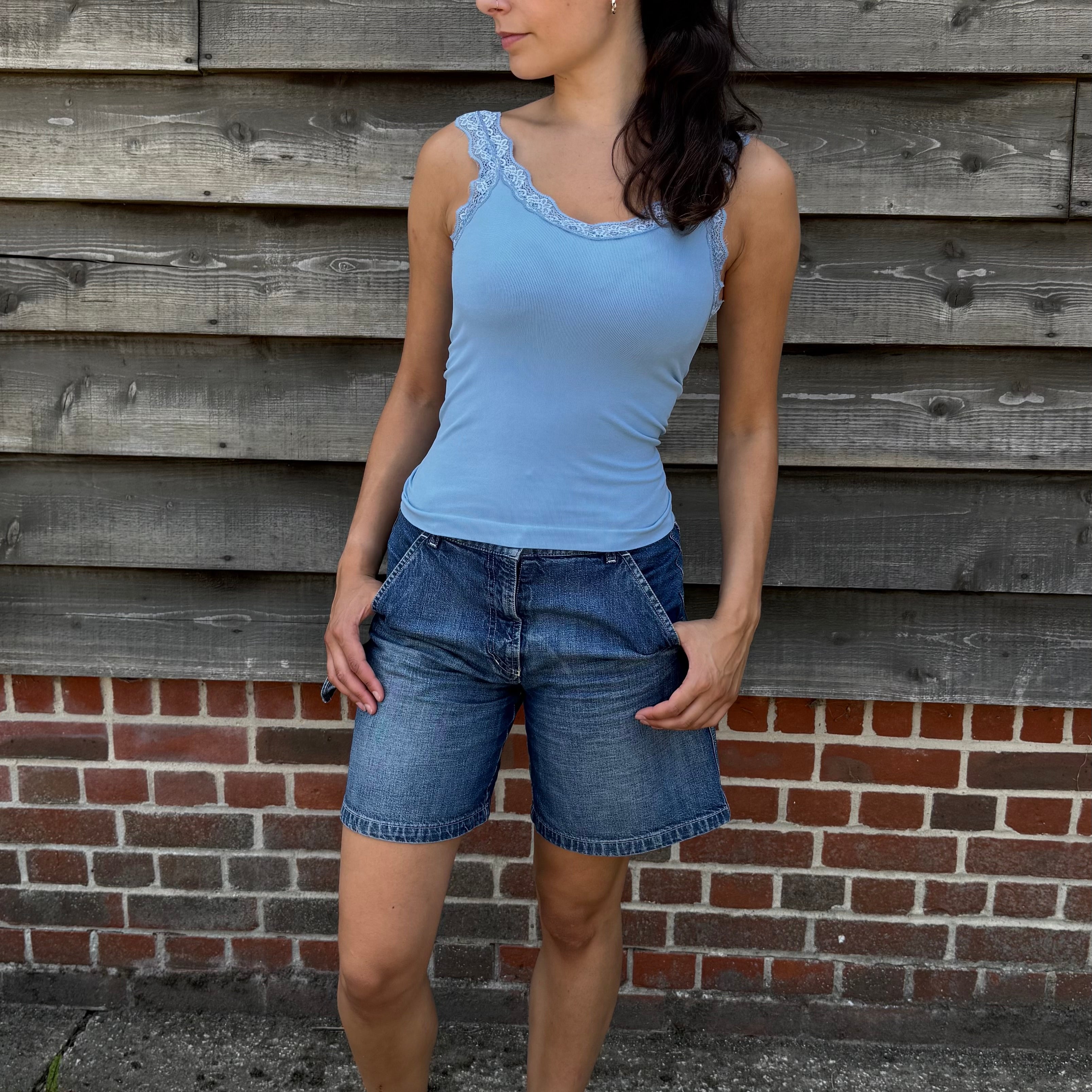 Woman wearing a blue tank top and denim shorts standing against a wooden wall.