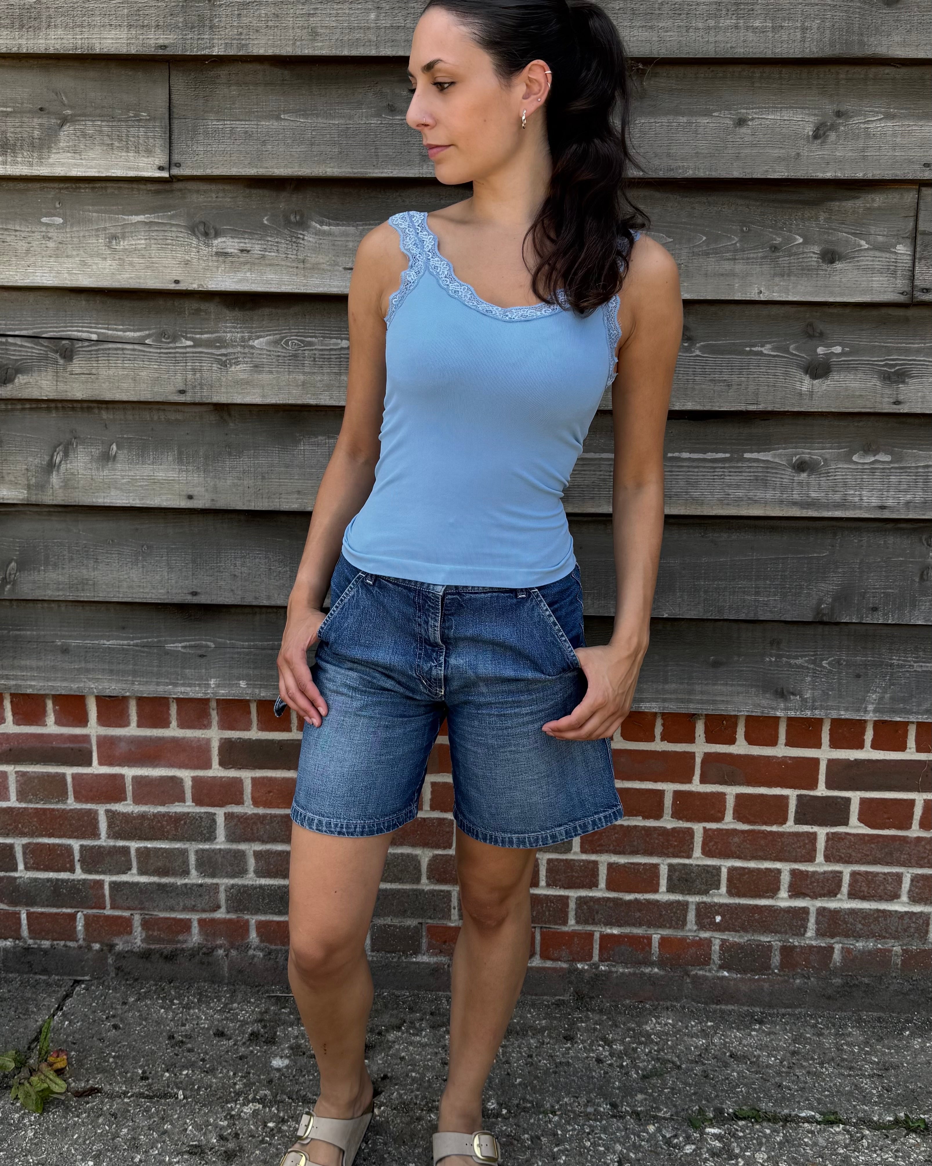 Woman wearing a blue tank top and denim shorts standing against a wooden wall.