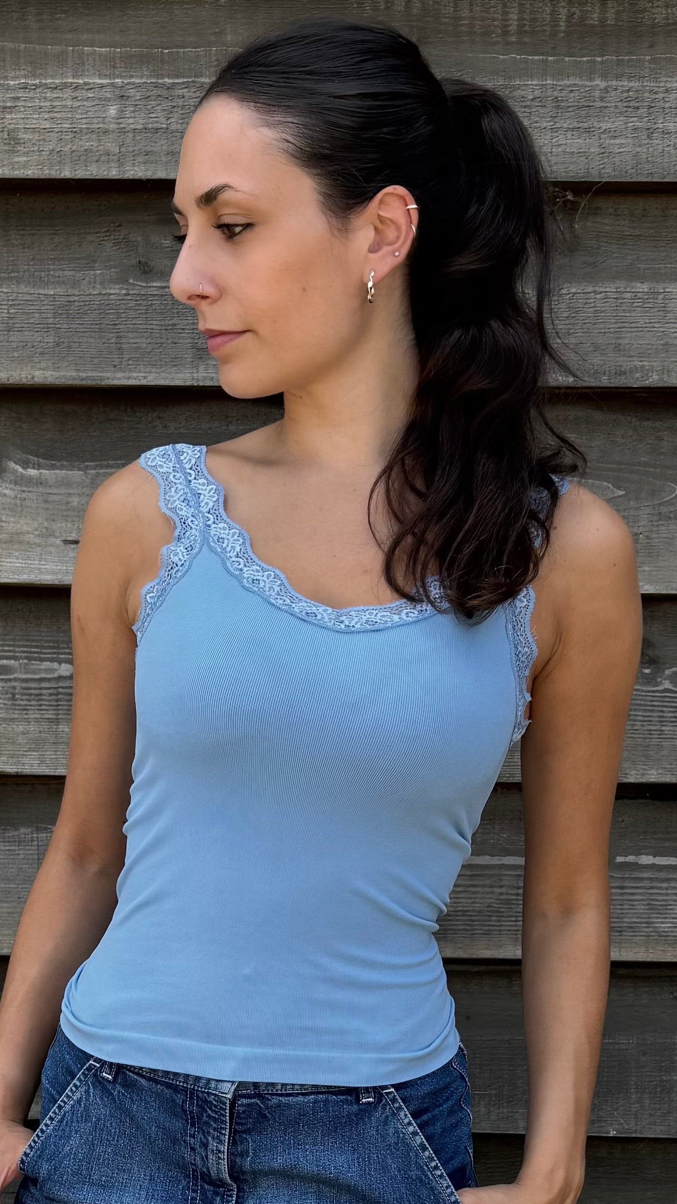 Woman wearing a blue tank top and denim shorts standing against a wooden wall.