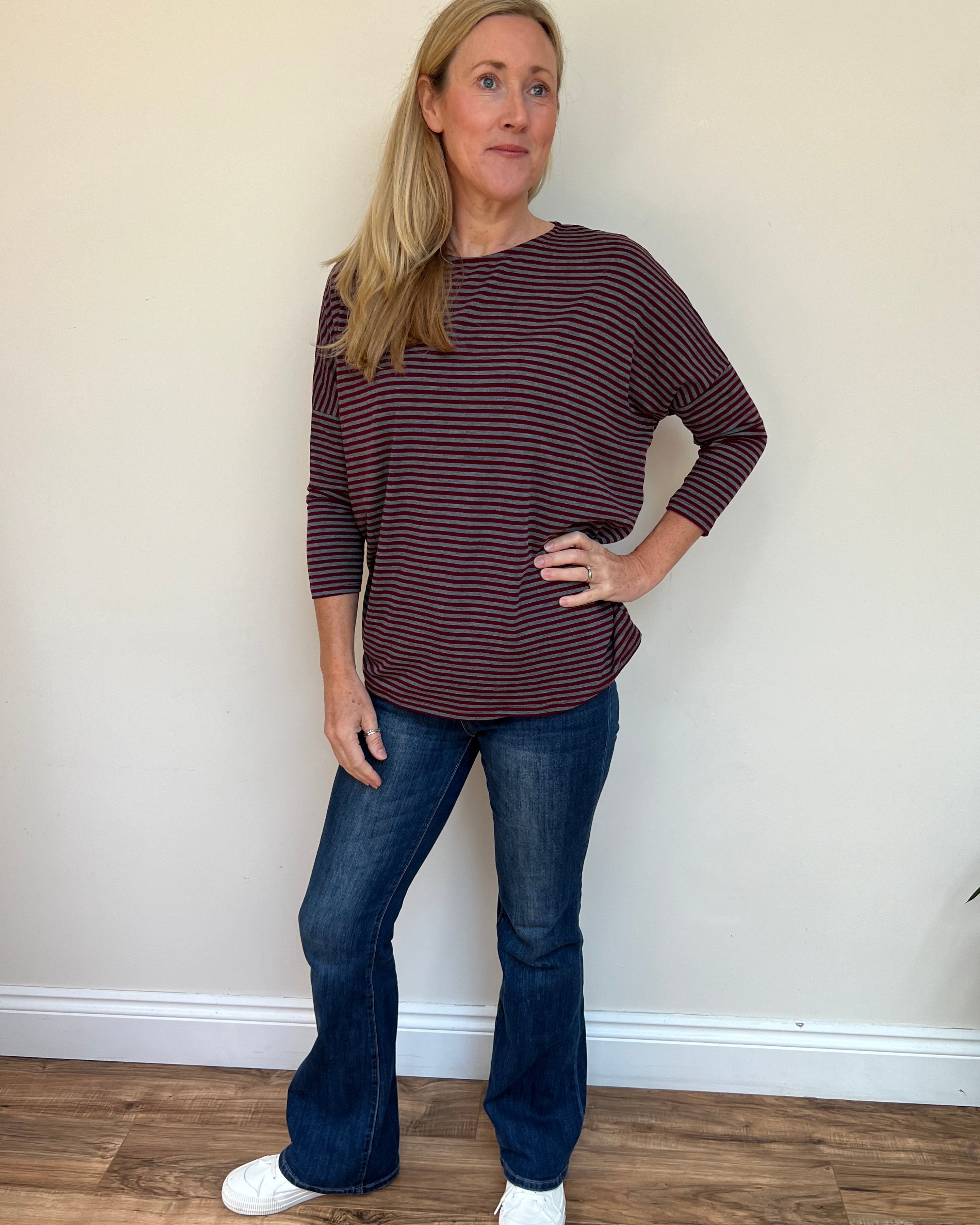 Woman wearing a maroon and white striped shirt and blue jeans standing against a plain wall.