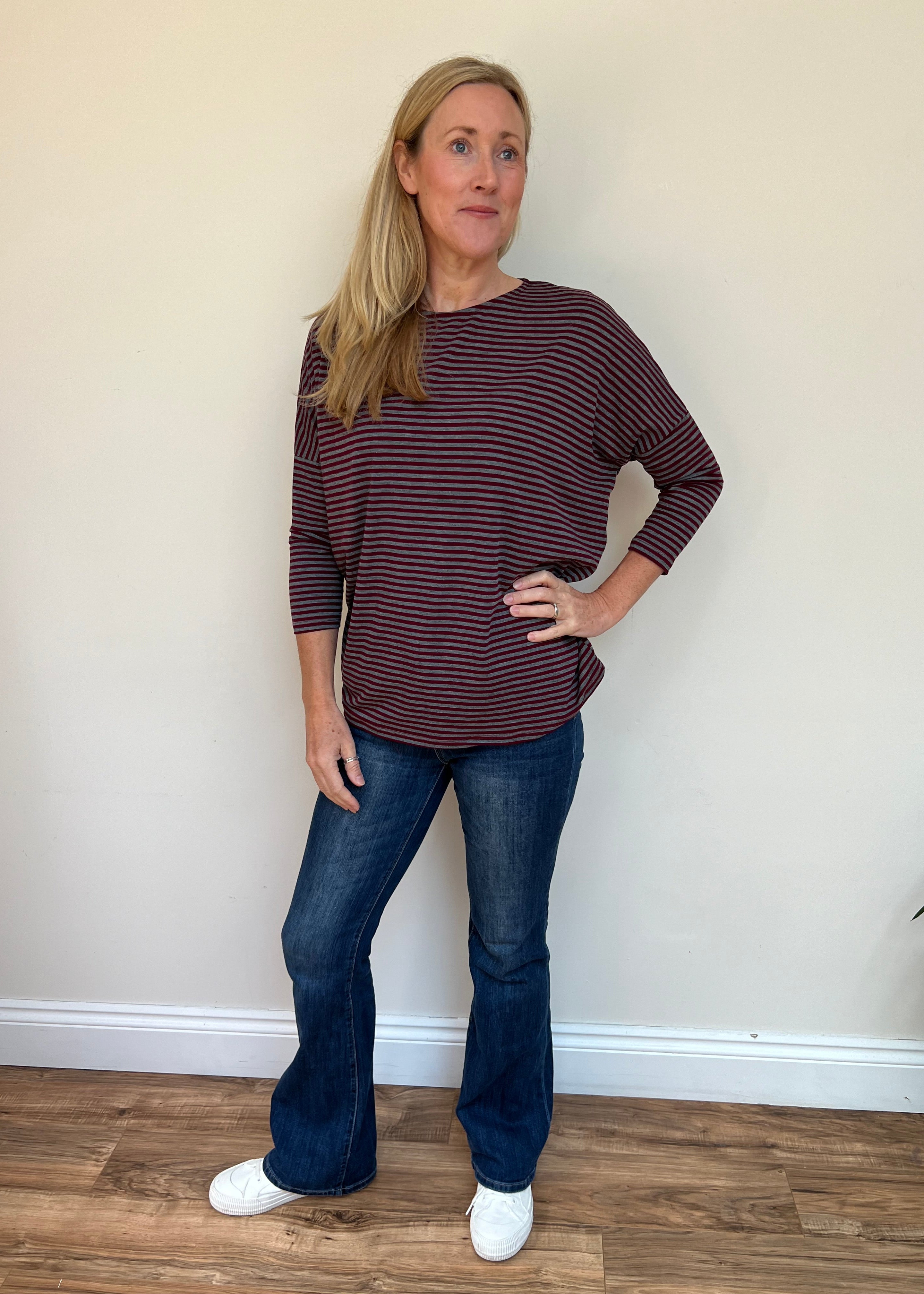 Woman wearing a maroon and white striped shirt and blue jeans standing against a plain wall.