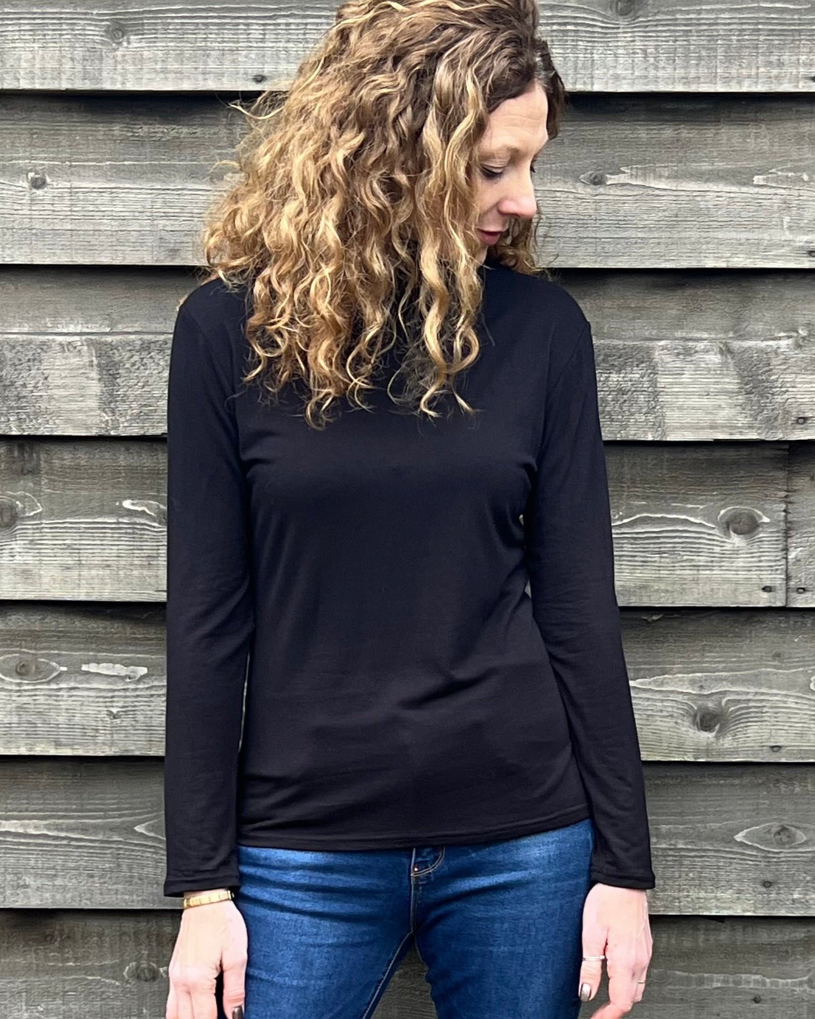Woman wearing a black long-sleeve top and blue jeans standing against a wooden wall.