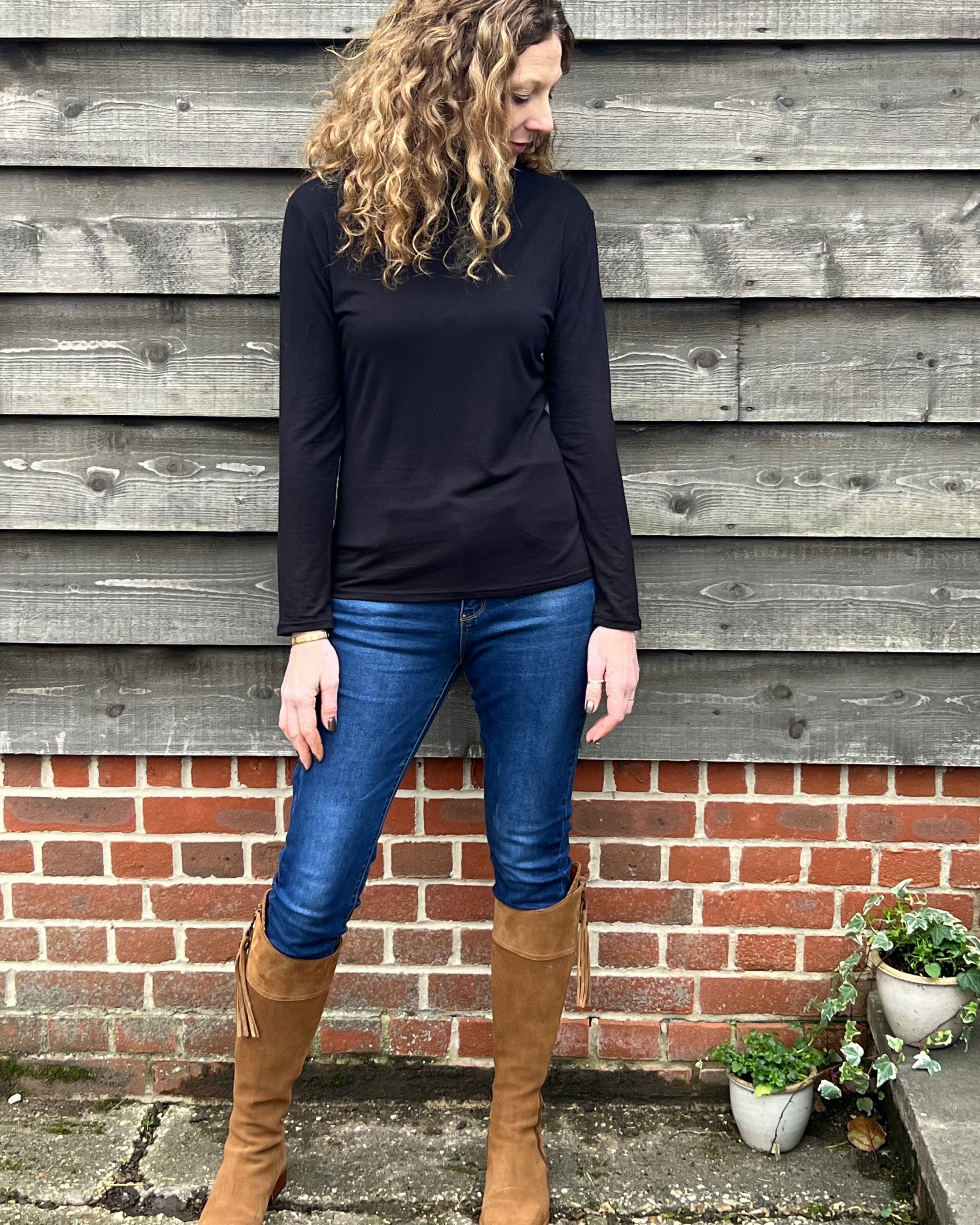 Woman wearing a black long-sleeve top and blue jeans standing against a wooden wall.