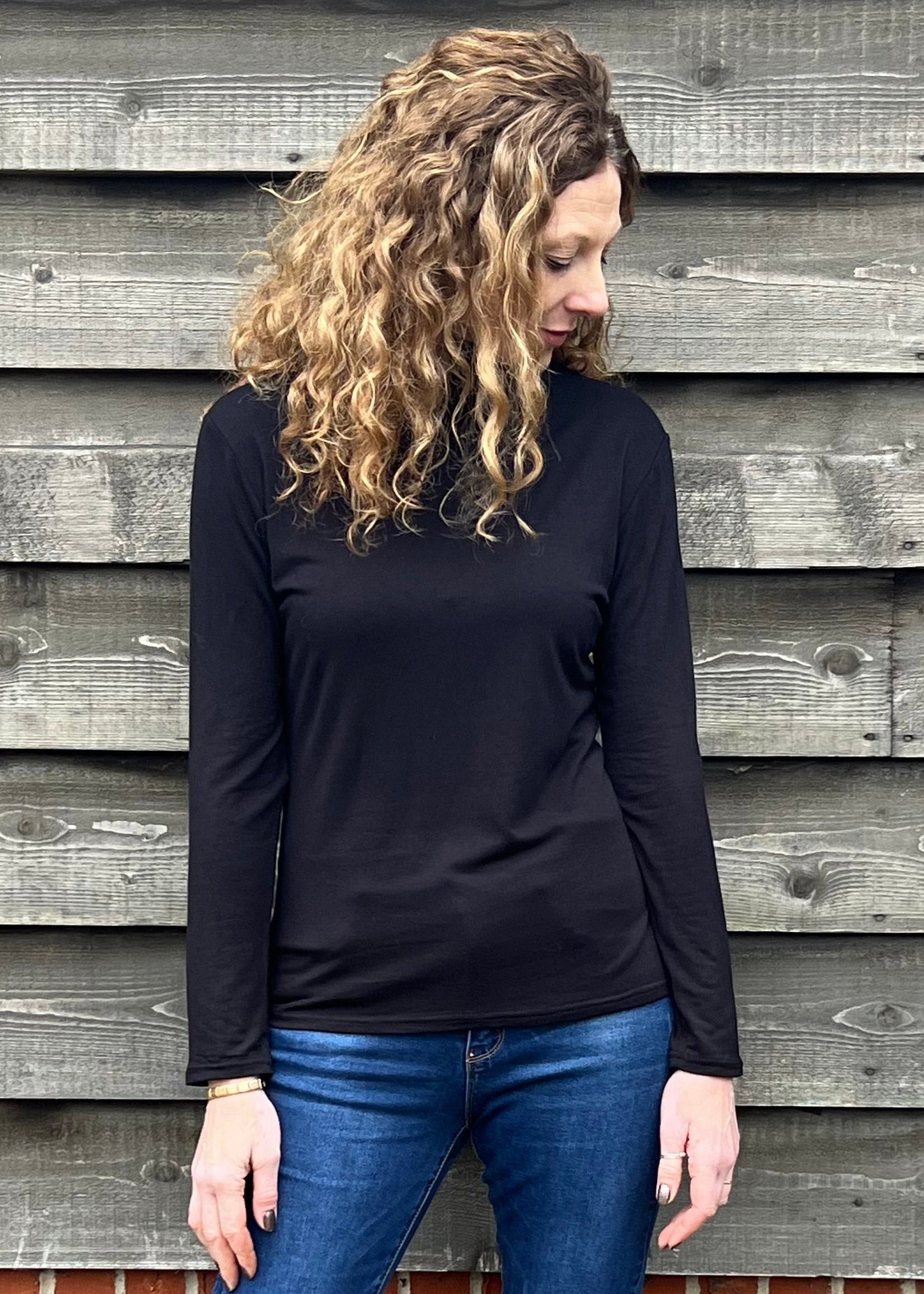 Woman wearing a black long-sleeve top and blue jeans standing against a wooden wall.