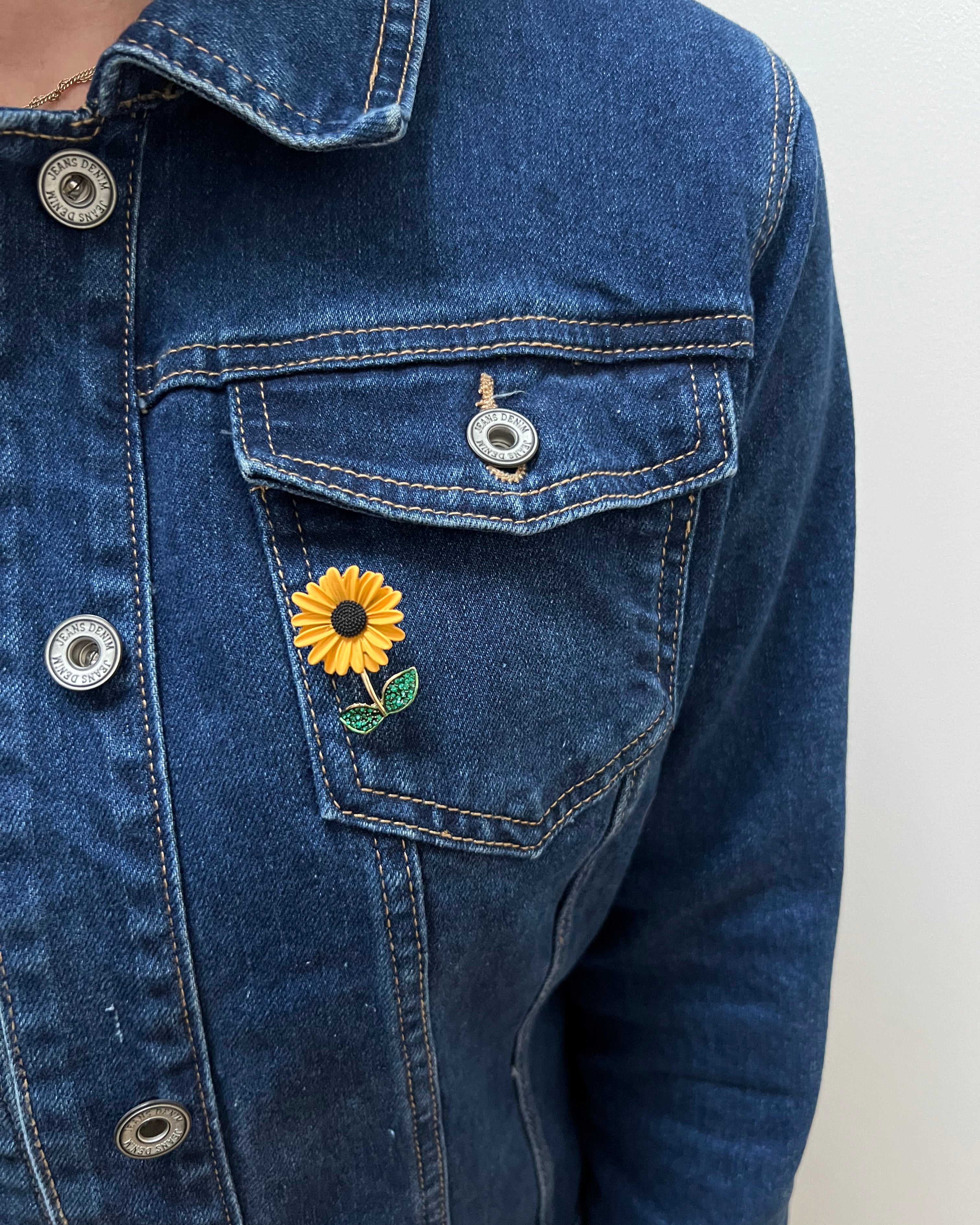A person wearing a blue denim jacket with a sunflower brooch on the lapel.