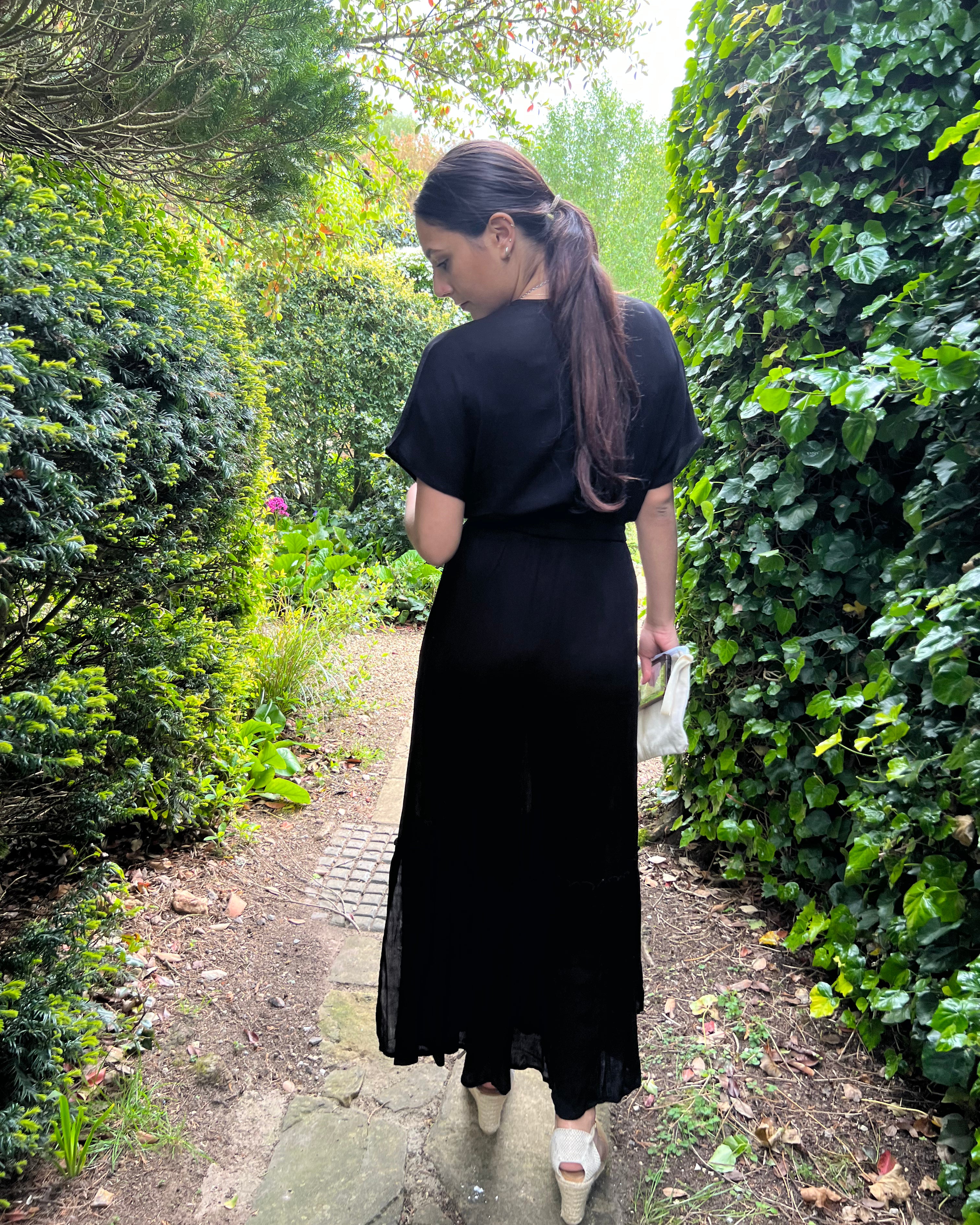 Person standing wearing a talia maxi dress in black by a door outside and trees.