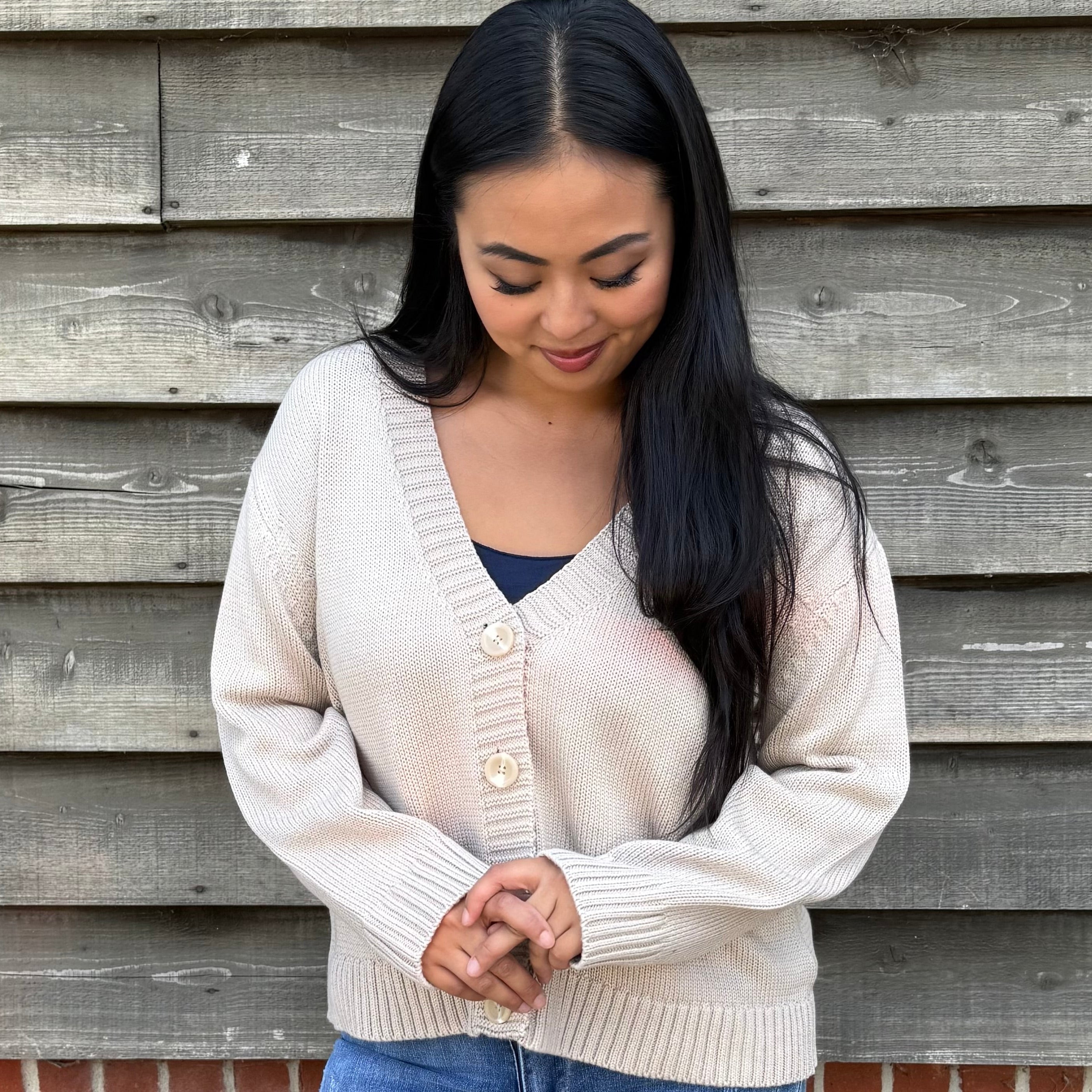 Woman wearing a beige cardigan and denim shorts standing against a wooden wall.