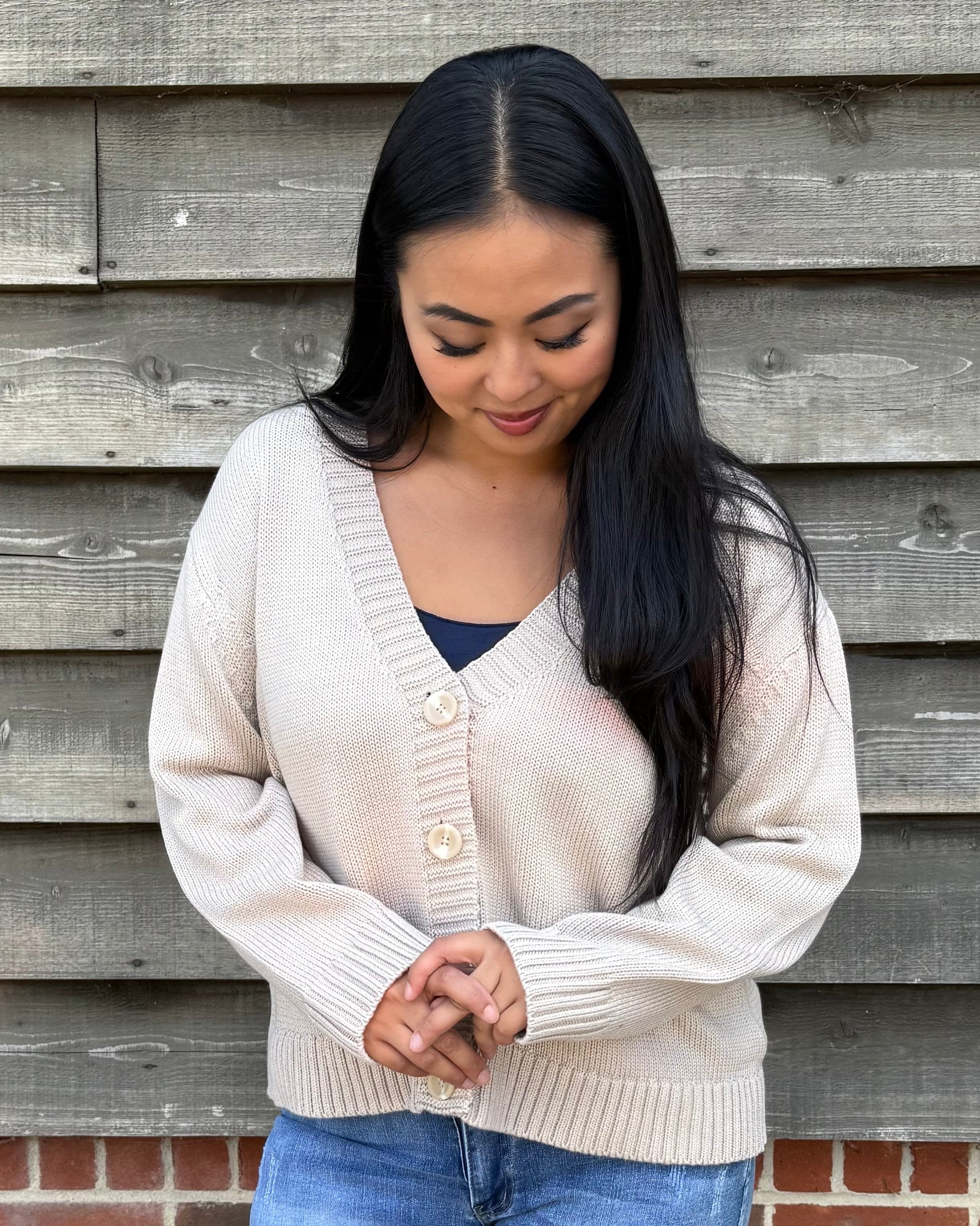 Woman wearing a beige cardigan and denim shorts standing against a wooden wall.