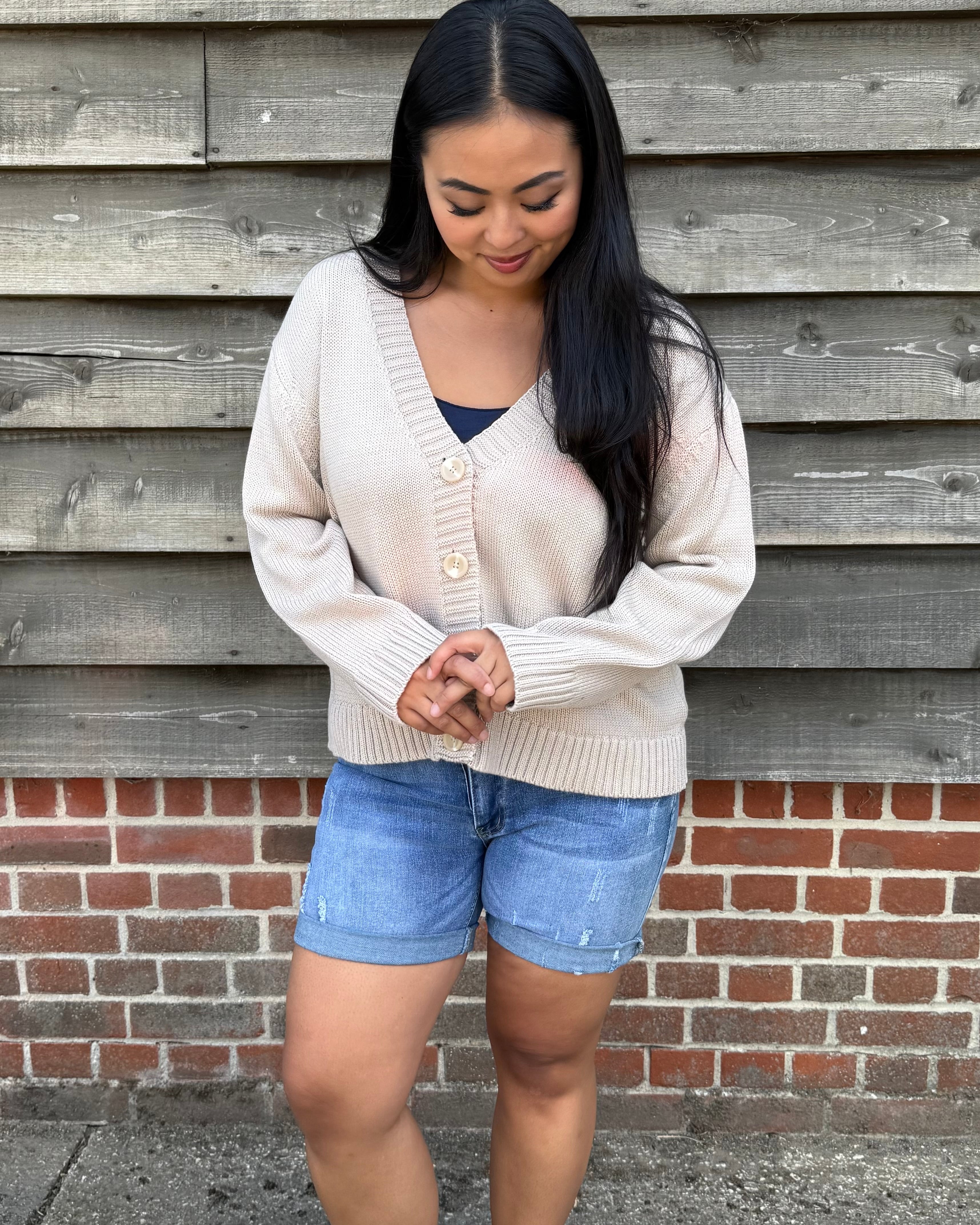 Woman wearing a beige cardigan and denim shorts standing against a wooden wall.